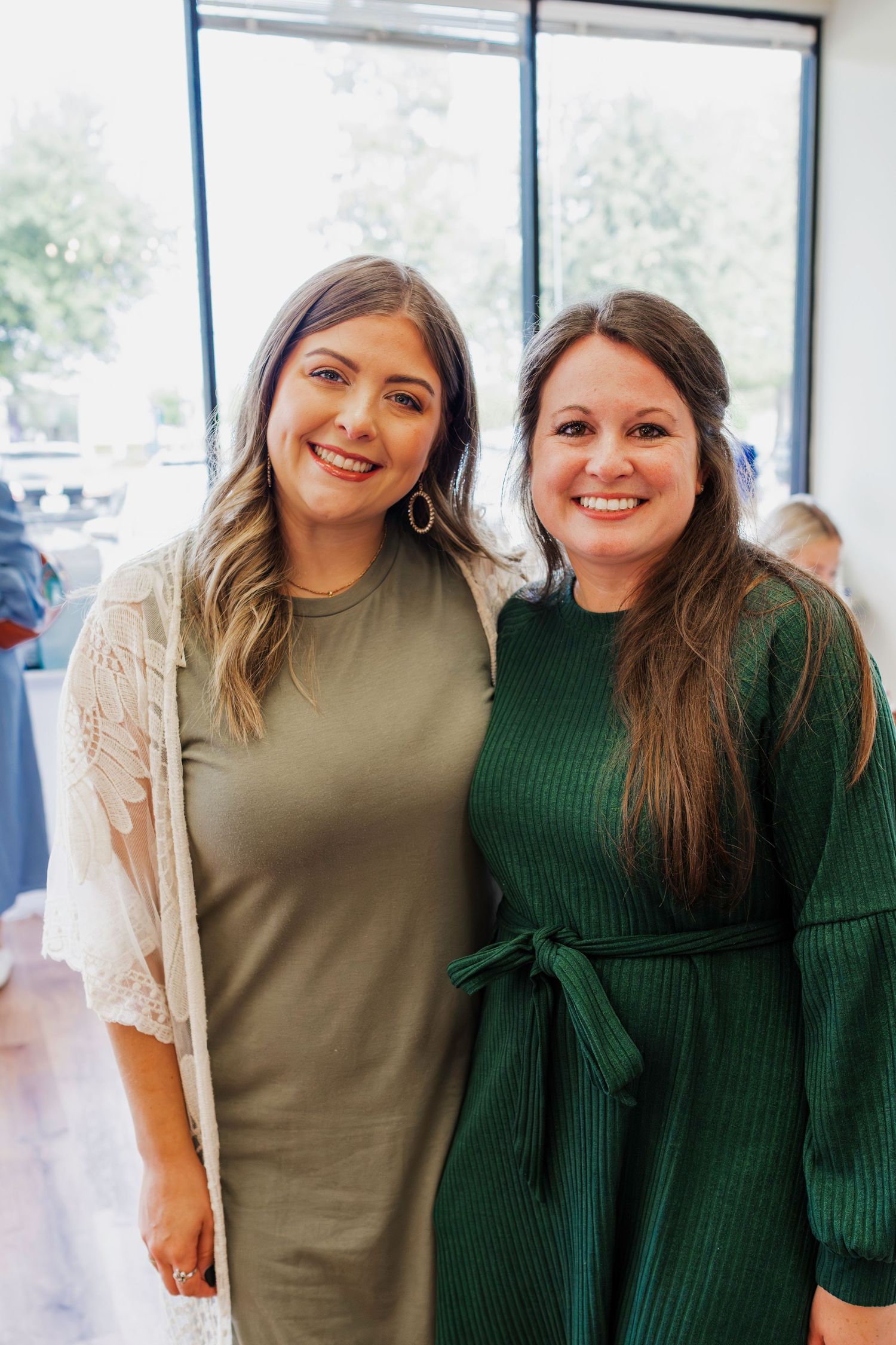 Two friends pose together at celebration wearing earth-toned and green dresses.