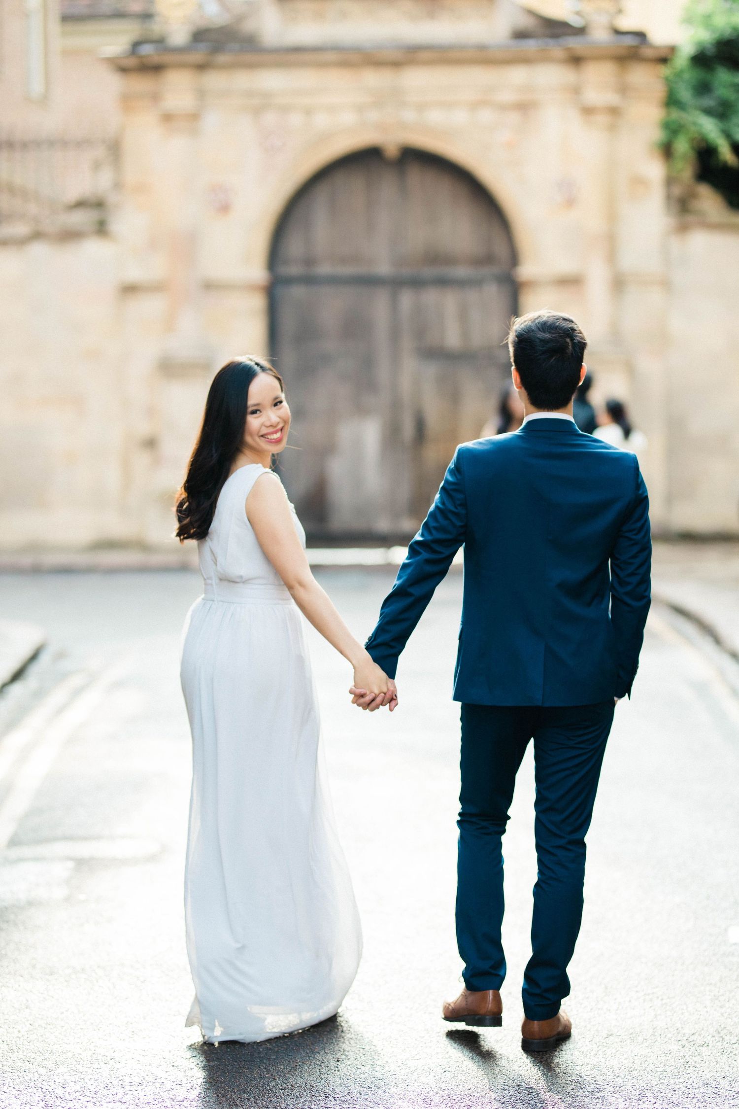 Natural engagement shoot moment with bride-to-be looking over shoulder at fiancé while strolling in Cambridge
