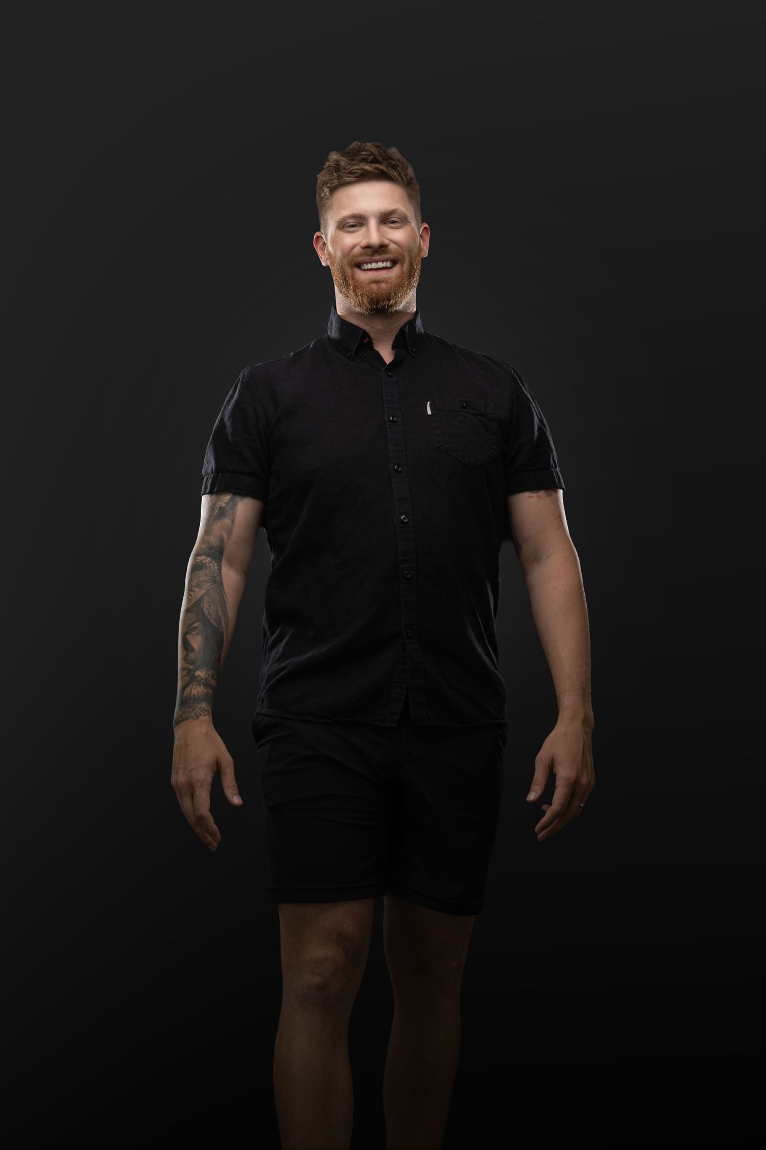 Smiling man with red beard and tattoo wearing black shirt and shorts against dark background.
