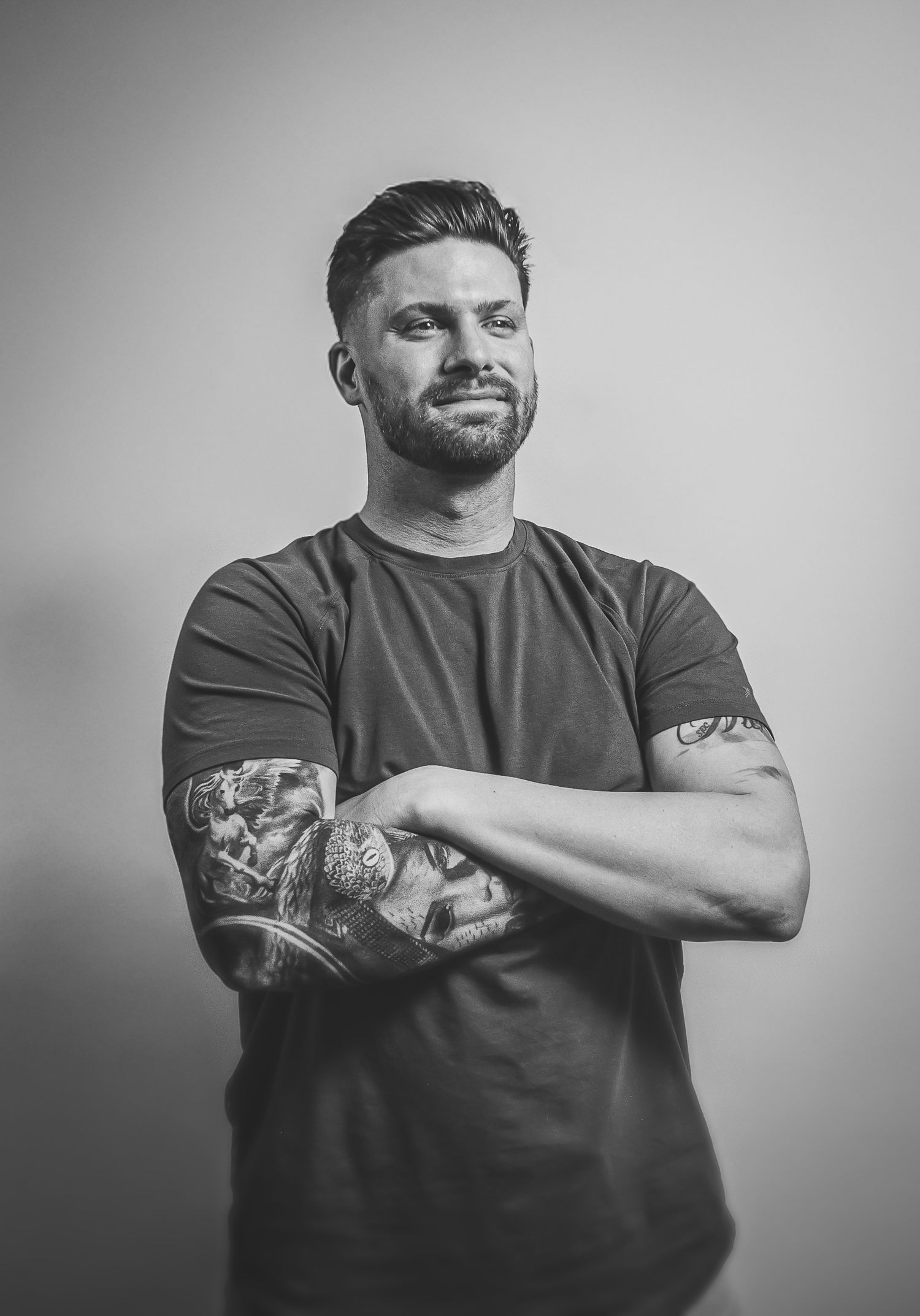 Black and white portrait of Roman Smith of Roman Thomas Media in t-shirt with sleeve tattoo against plain backdrop.