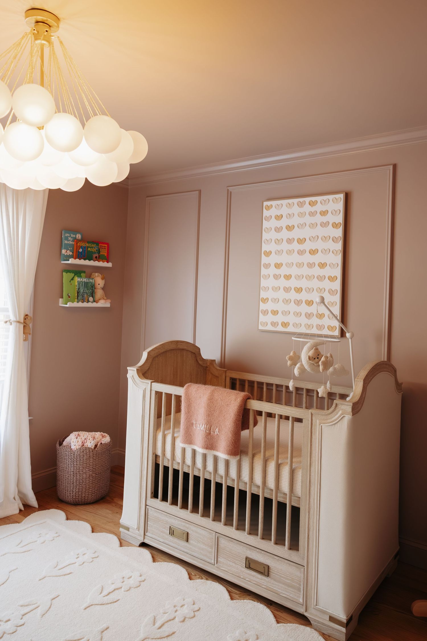 Elegant nursery with white crib, pink blanket, globe chandelier, wall art, and book shelves in a warm taupe room.