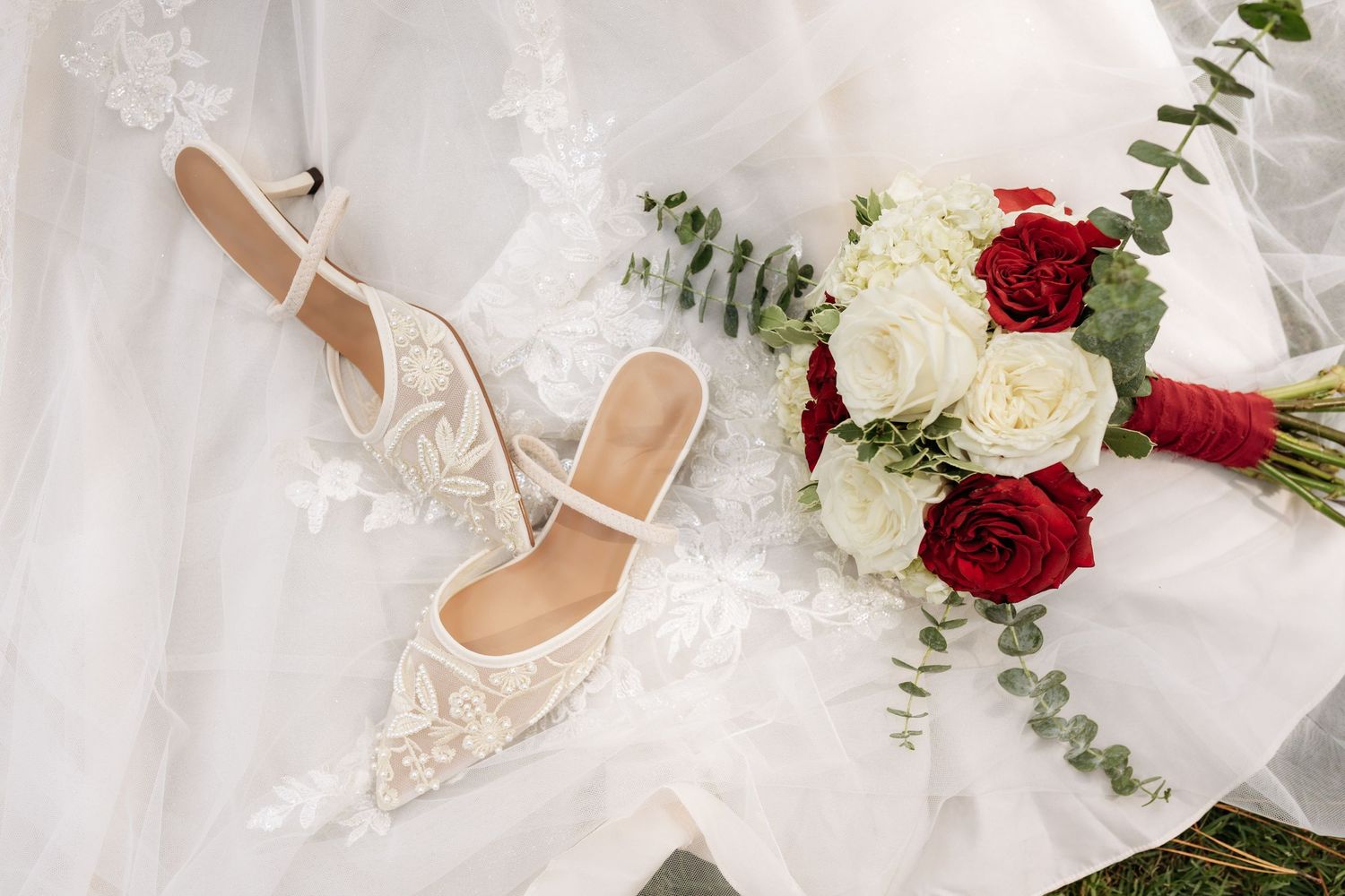 Elegant bridal flat shoes with lace detail beside a red and white rose bouquet on a white wedding dress.