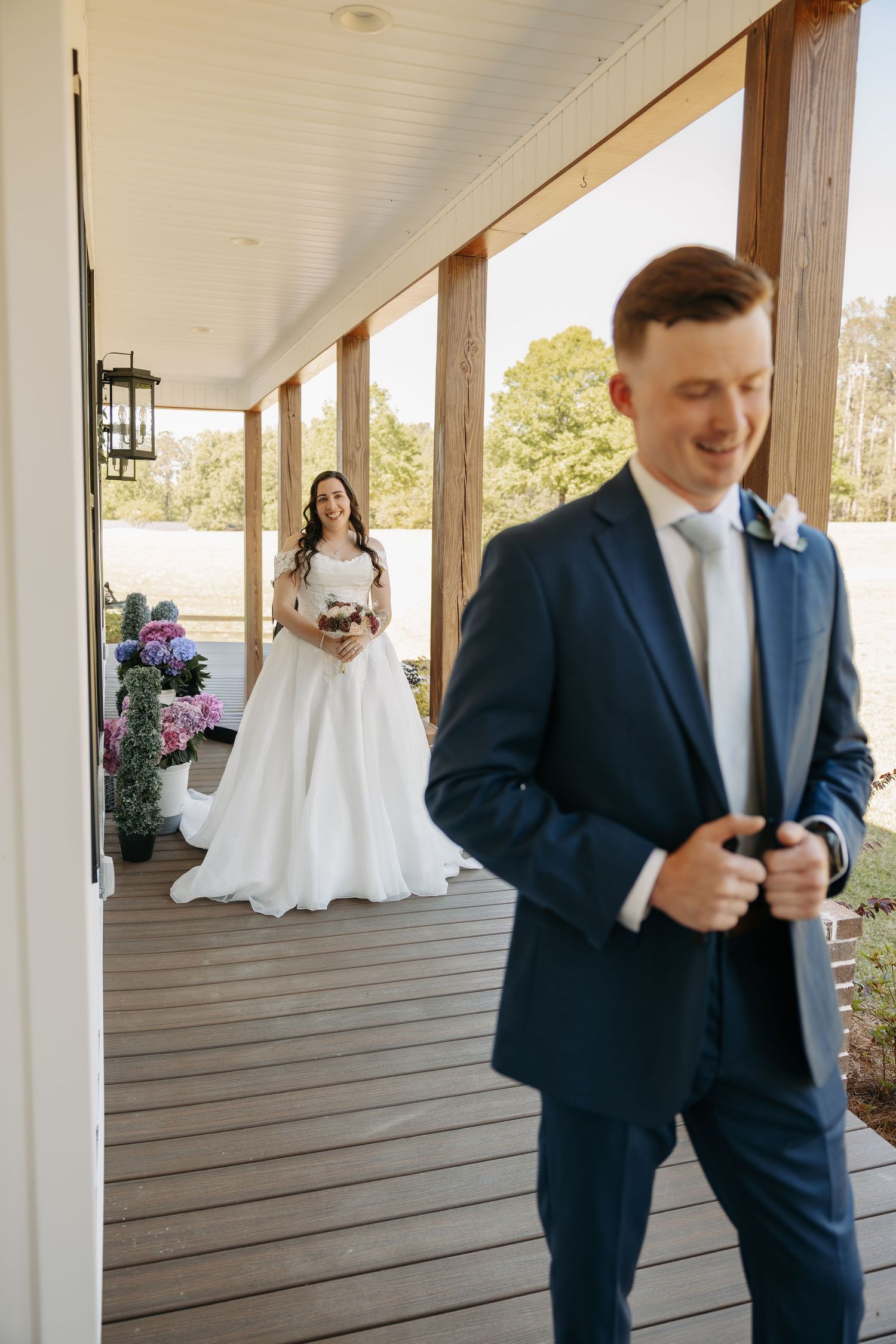 Bride in white gown holds bouquet on porch while groom in navy suit smiles in foreground during first look.