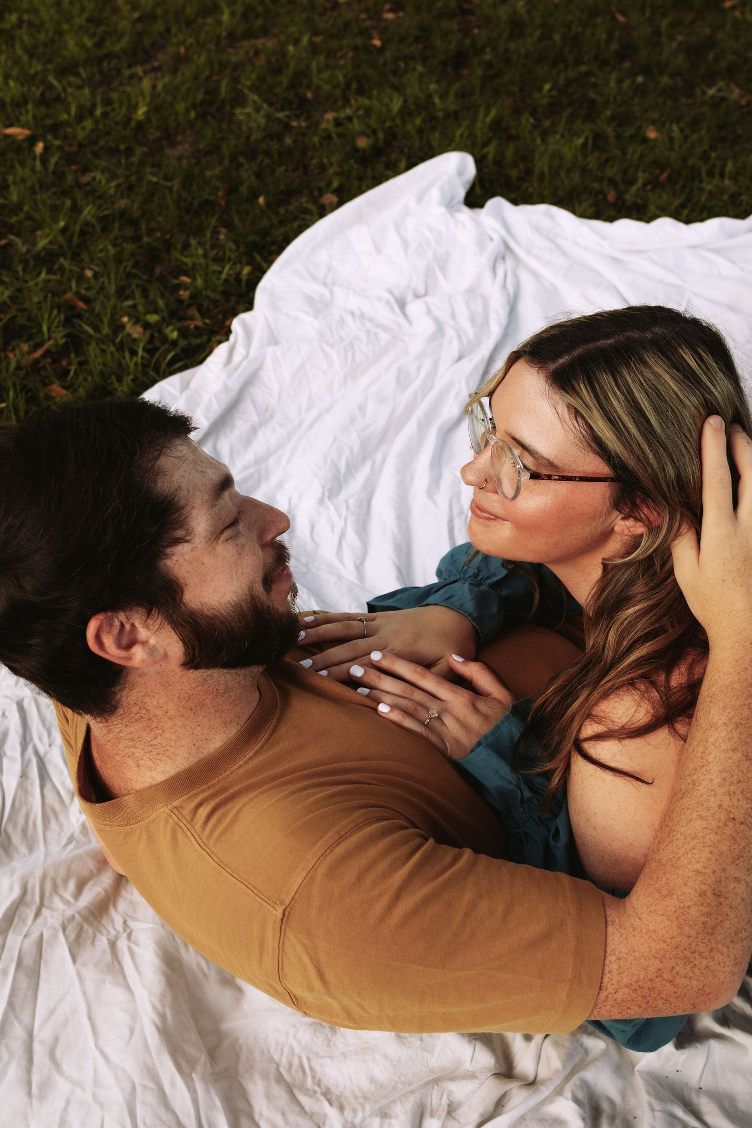 A couple lying on a white blanket in the grass, gazing at each other romantically during a sunny outdoor picnic.