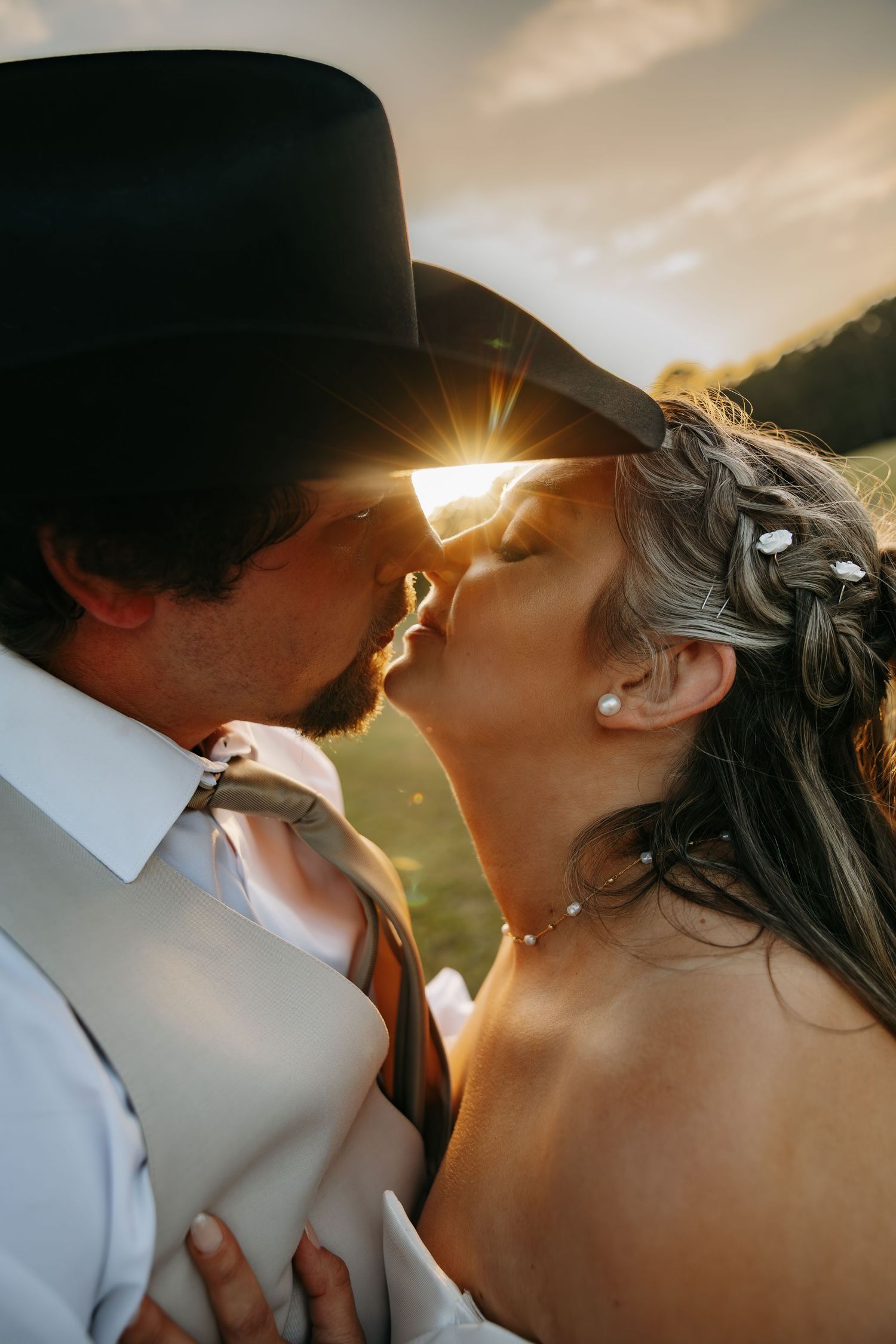 Romantic close-up of bride and groom kissing at sunset with golden light flaring through cowboy hat.