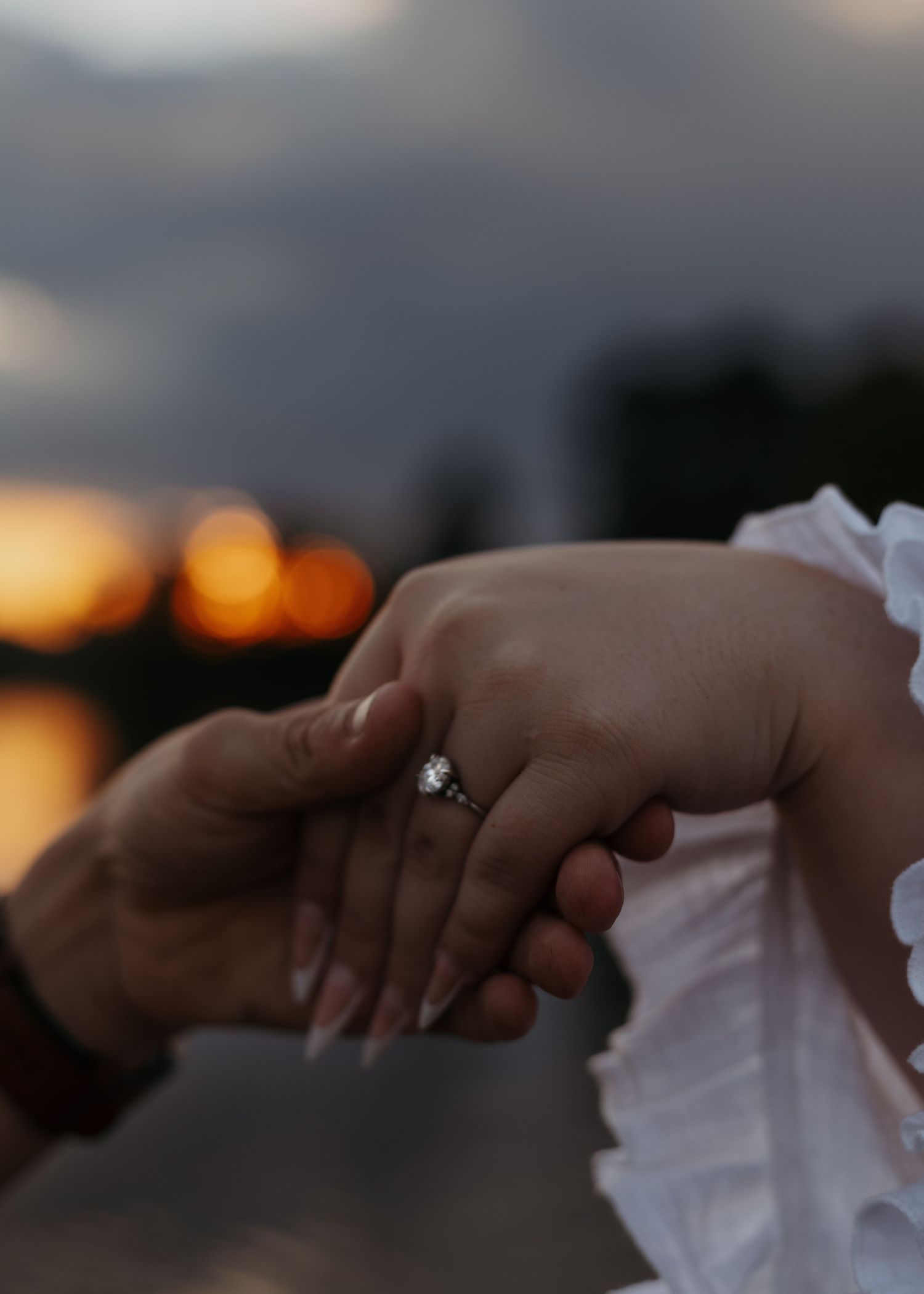 Two hands holding gently at sunset, one adorned with a sparkling diamond engagement ring, with a warm golden bokeh background.