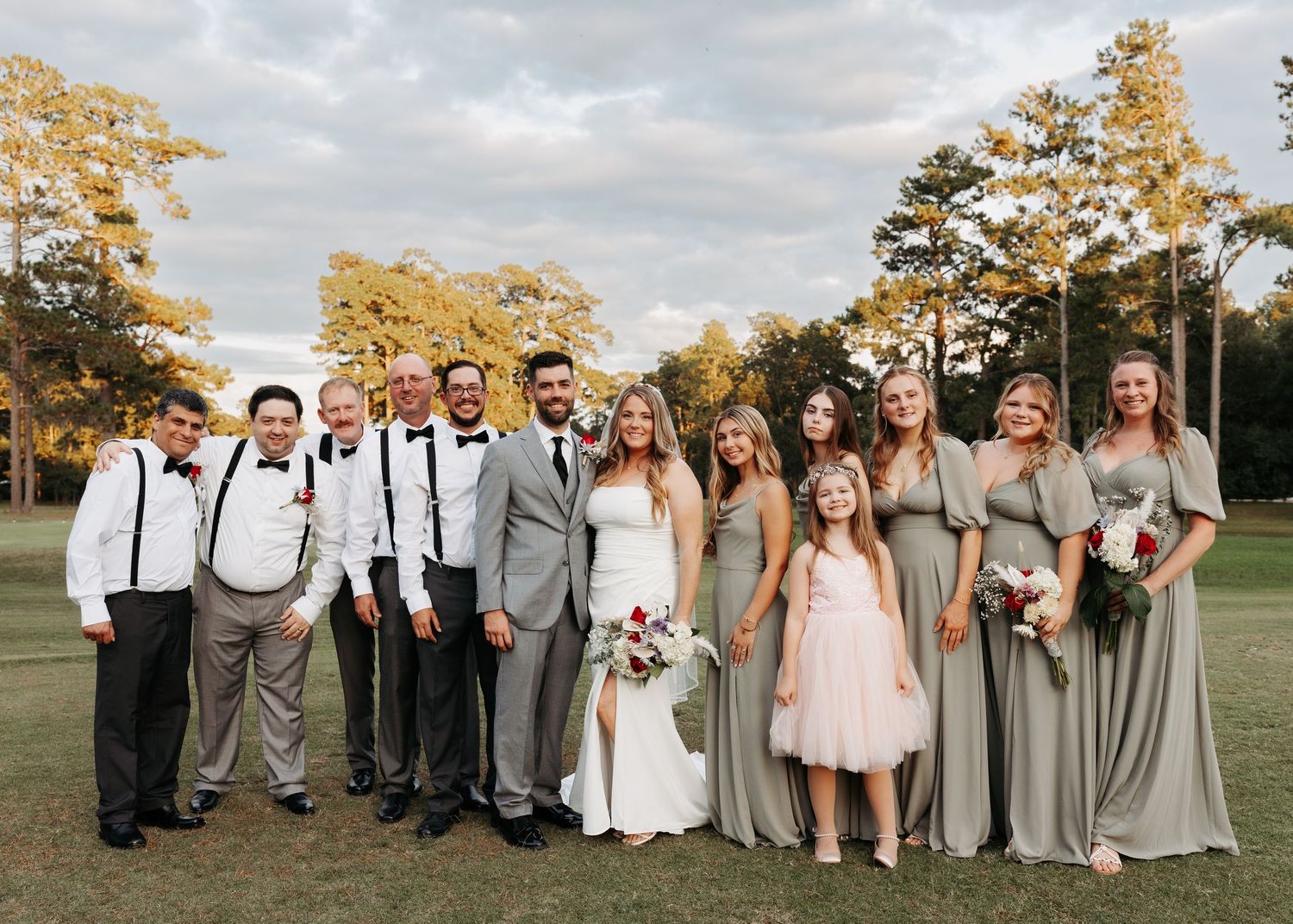 Wedding party posing outdoors on green lawn, bride in white gown, groom in gray suit, bridesmaids in sage green dresses.