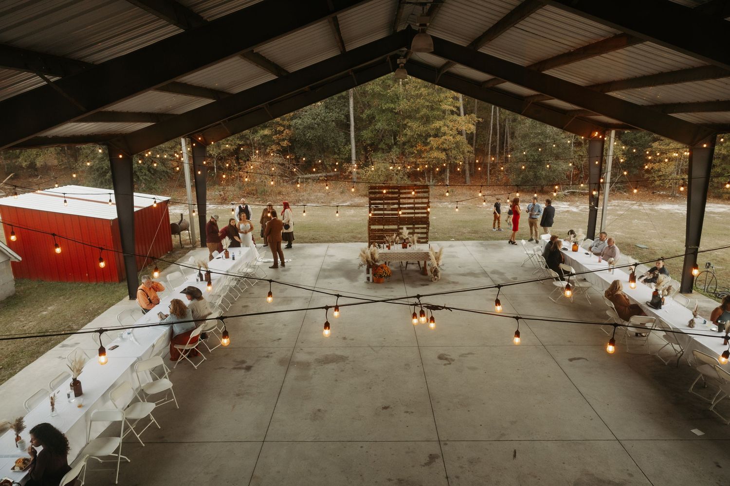 Aerial view of rustic outdoor wedding reception under pavilion with string lights and long tables.