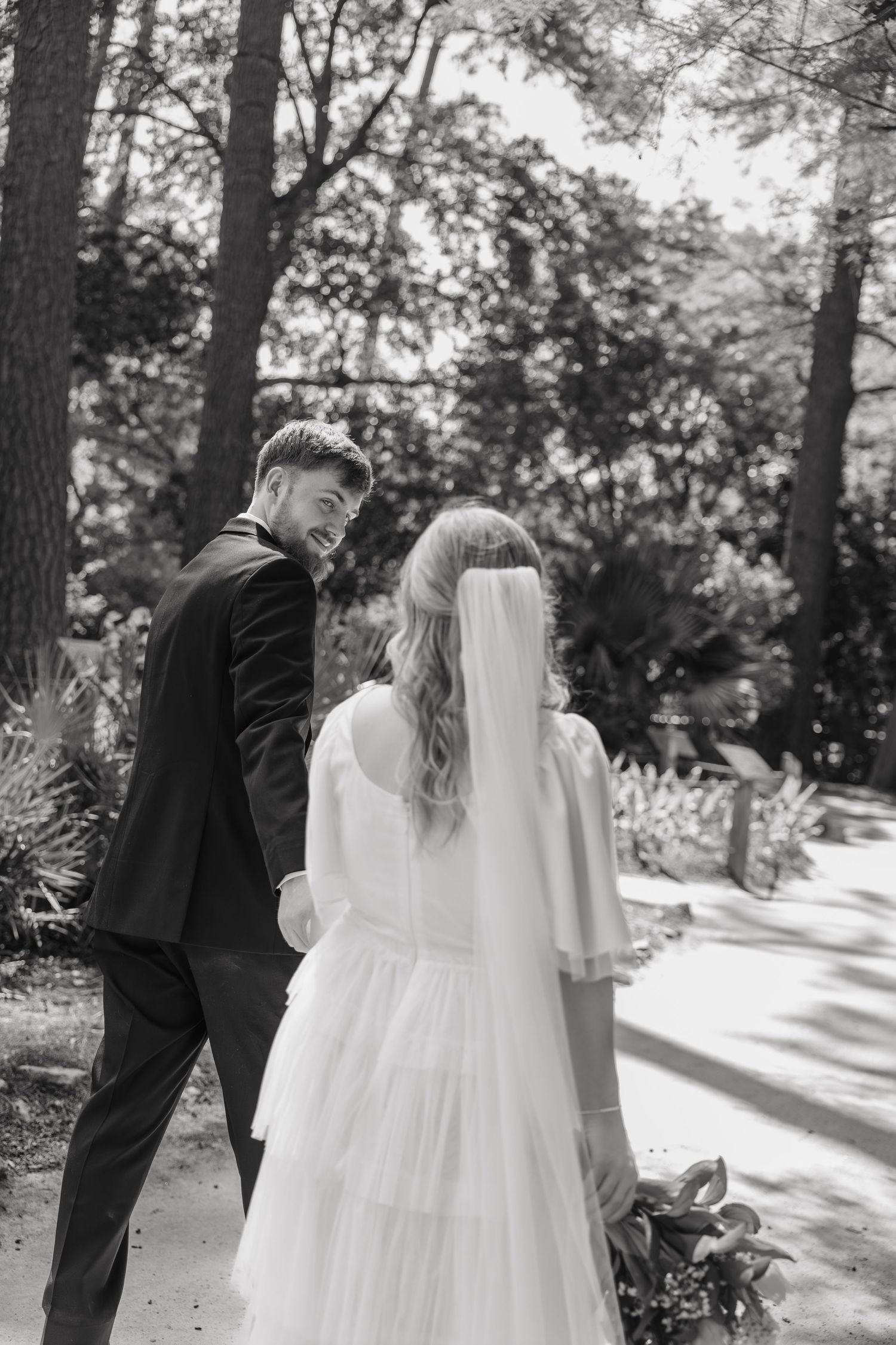 Black and white photo of smiling groom walking with bride in a garden path.