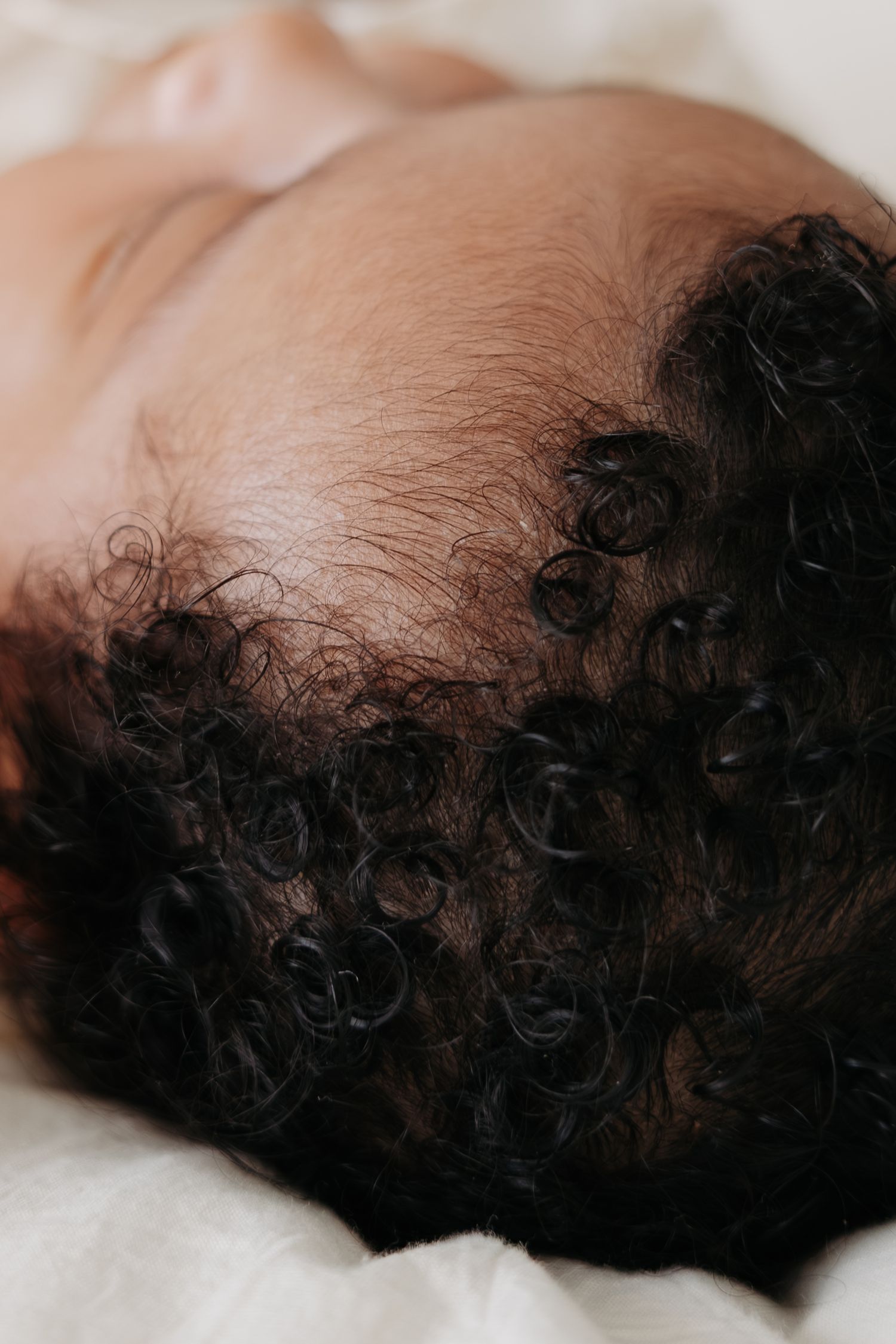 Close-up of a newborn baby's head with soft curly black hair against a white blanket.
