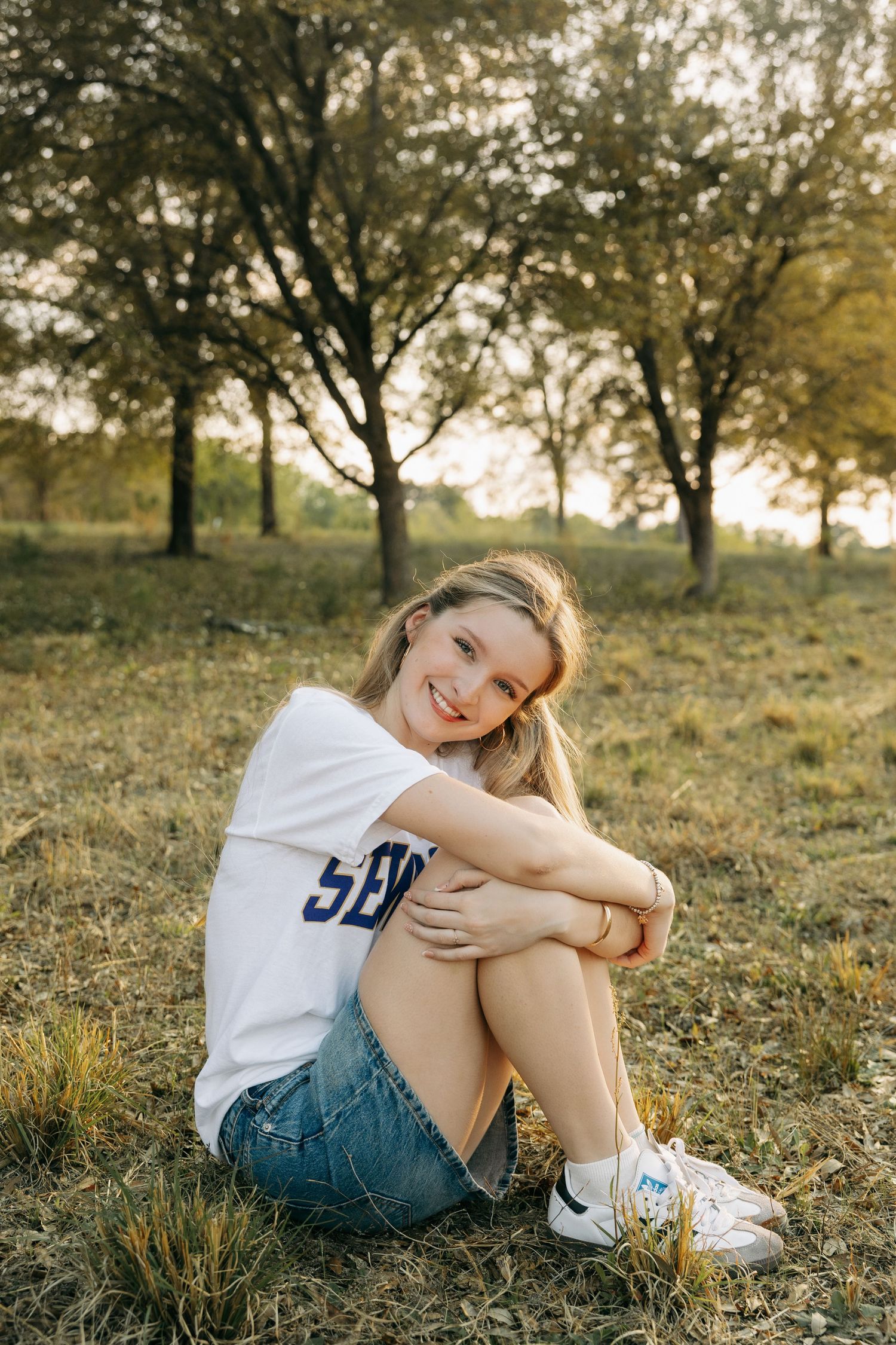 Young blonde woman sitting on grass in a park, wearing a white graphic tee and denim shorts, smiling cheerfully.