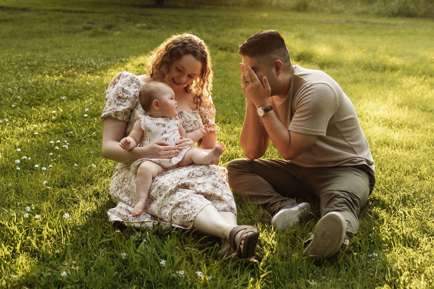 Young family sitting on green grass in sunlight, mother holding baby while father looks on adoringly.