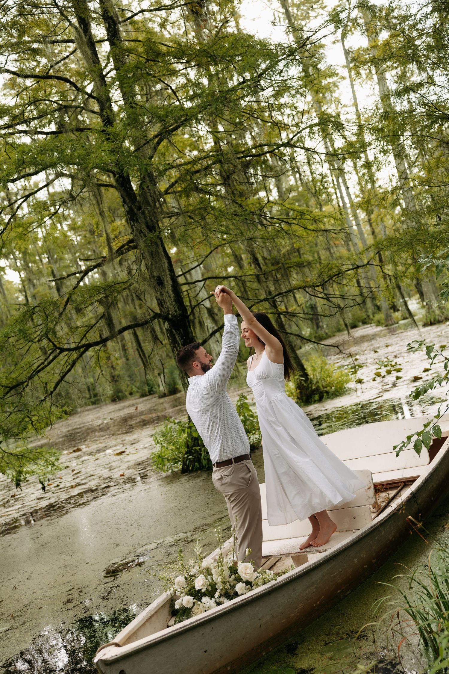 Groom twirling bride in white dress on a decorated boat surrounded by cypress trees.