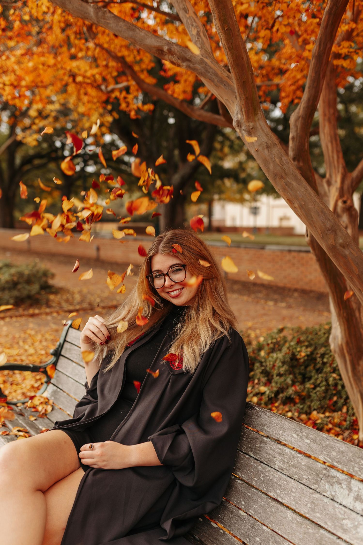 Autumn graduation photoshoot on wooden bench with orange leaves and trees in background.