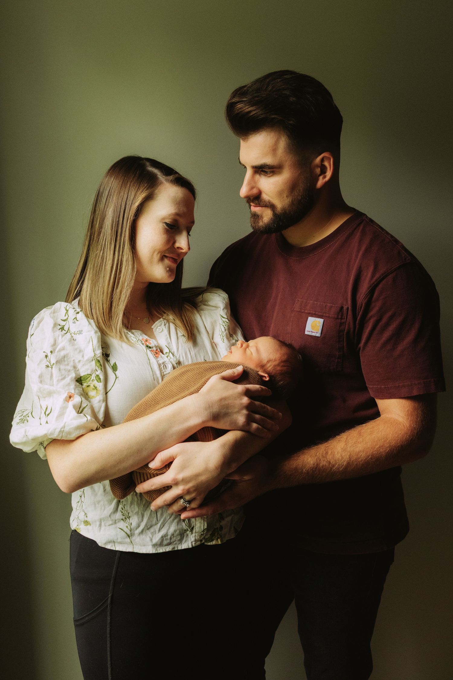 An intimate family photography series in moody lighting shows tender moments between parents and their newborn baby.