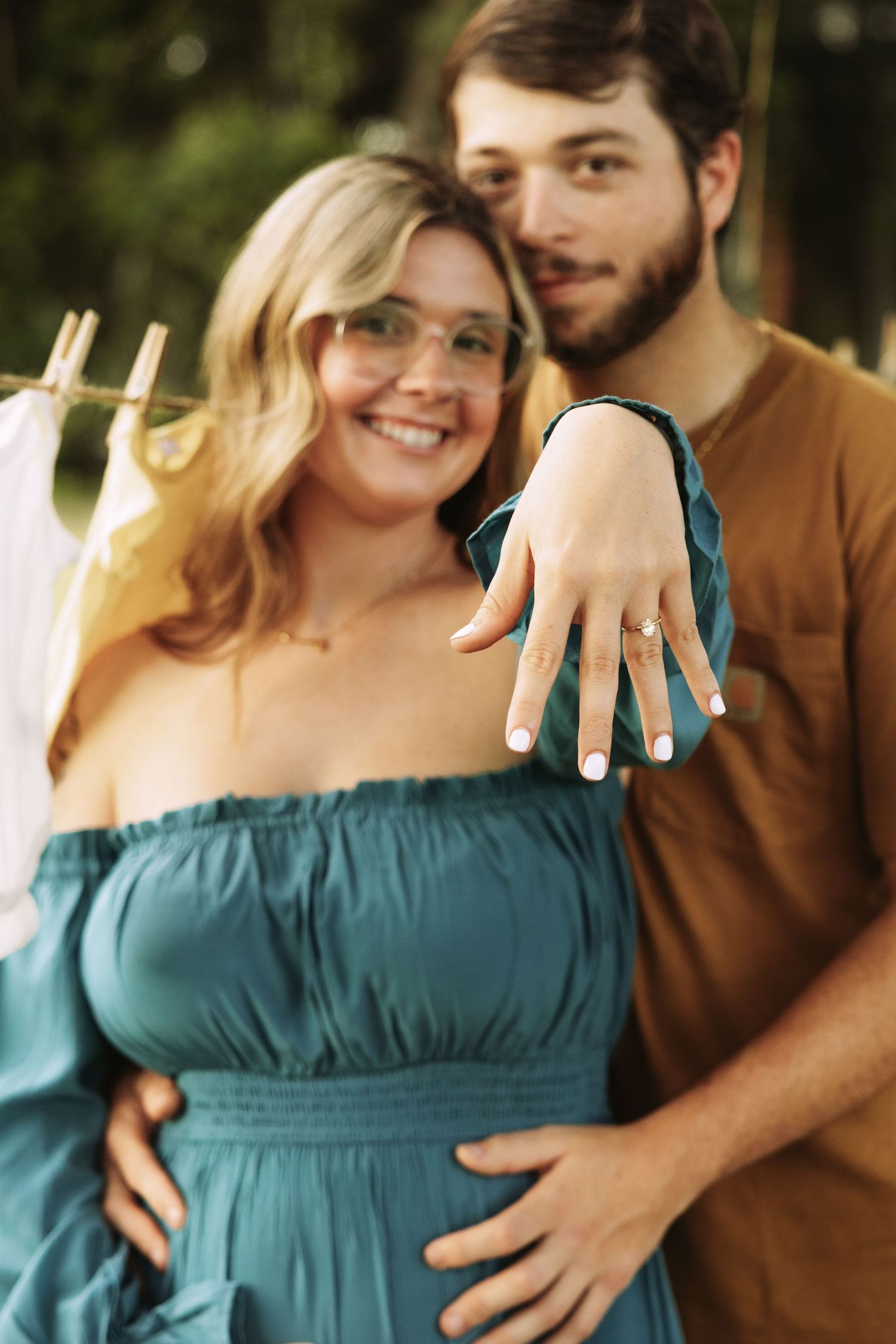 A couple shows off their engagement ring in a romantic outdoor setting with the person in teal dress displaying their hand.