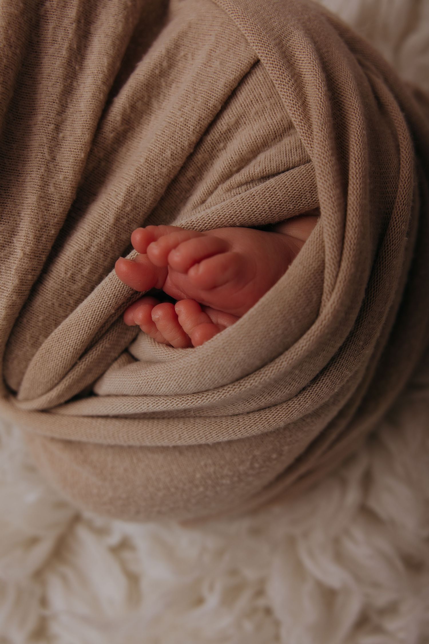 Tiny baby feet peek out from beneath a soft brown blanket in a tender newborn photography series.