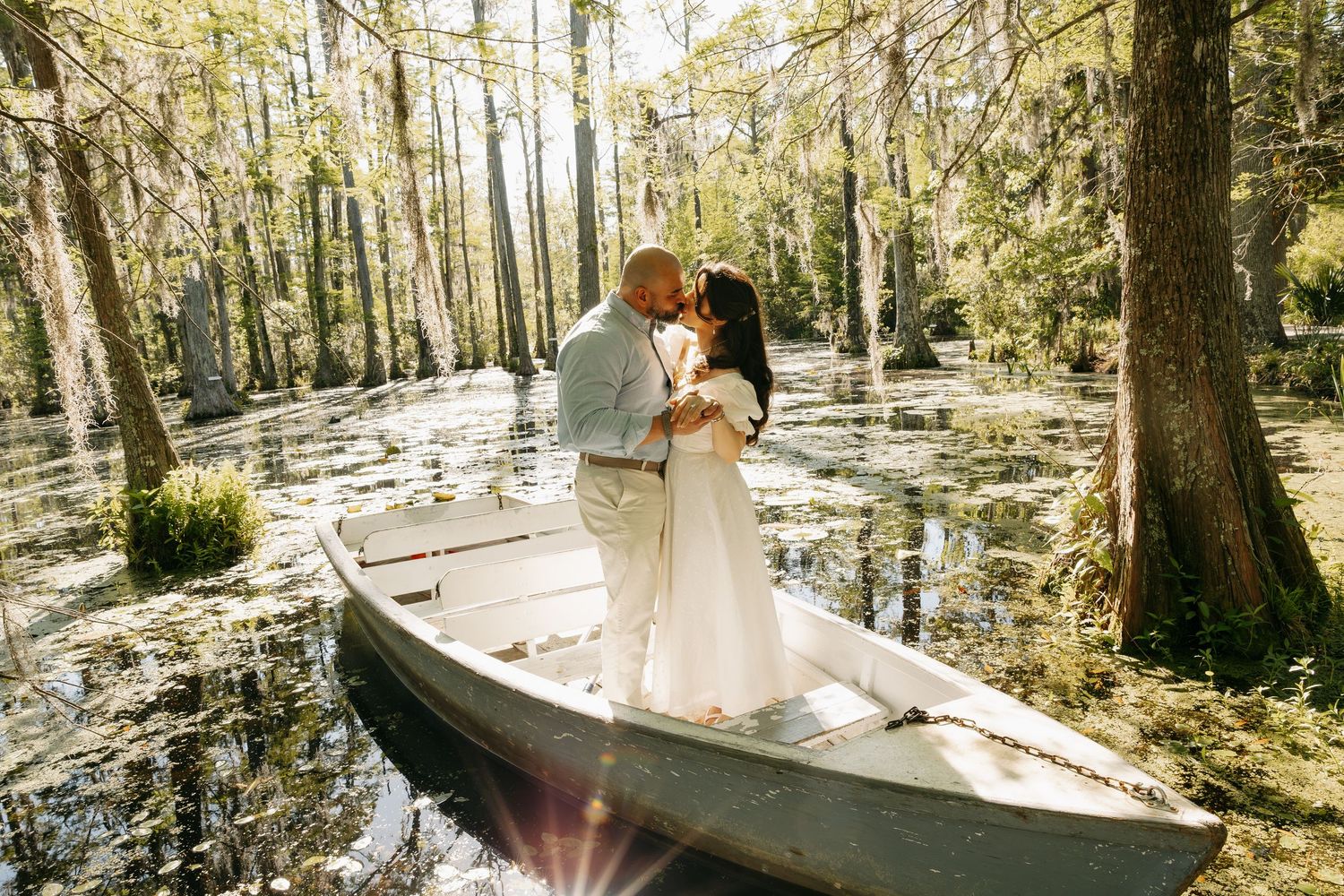 Couple sharing a romantic kiss in a white rowboat surrounded by a sunlit cypress swamp forest.