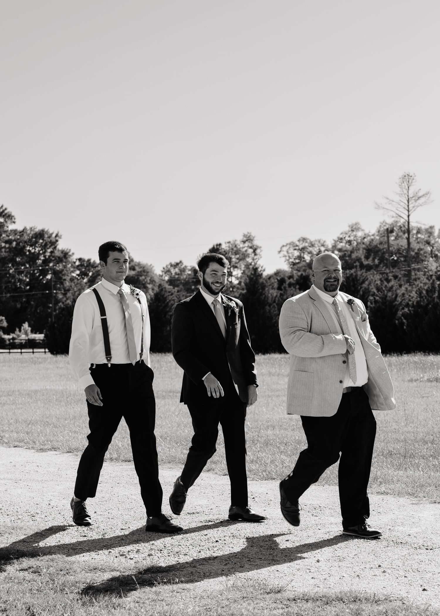 Black and white photo of groom and groomsmen walking outdoors in suits and suspenders.
