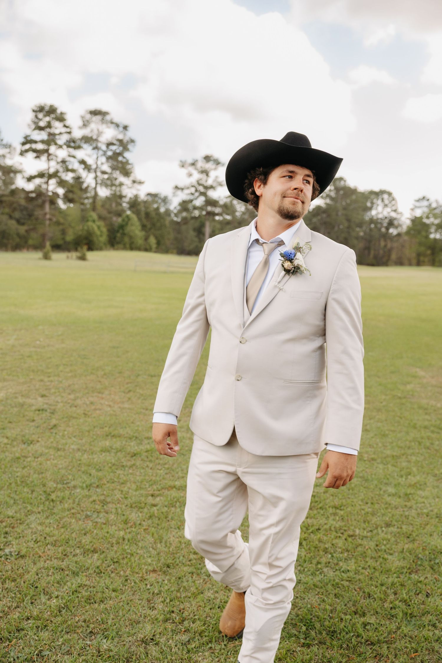 Handsome groom in cream suit and black cowboy hat walking across green field with boutonniere on lapel.
