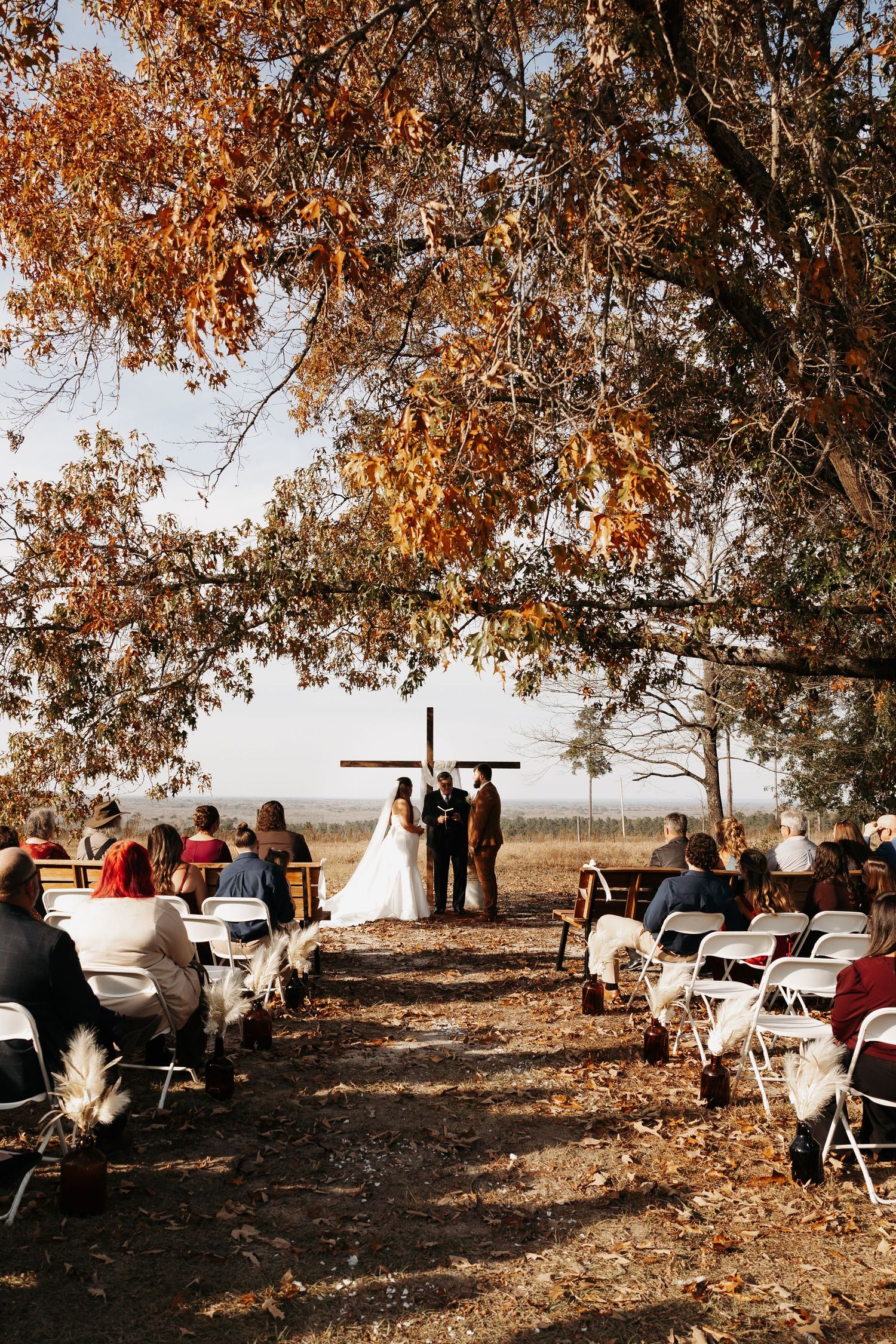Outdoor fall wedding ceremony under golden autumn trees with couple at wooden cross altar.