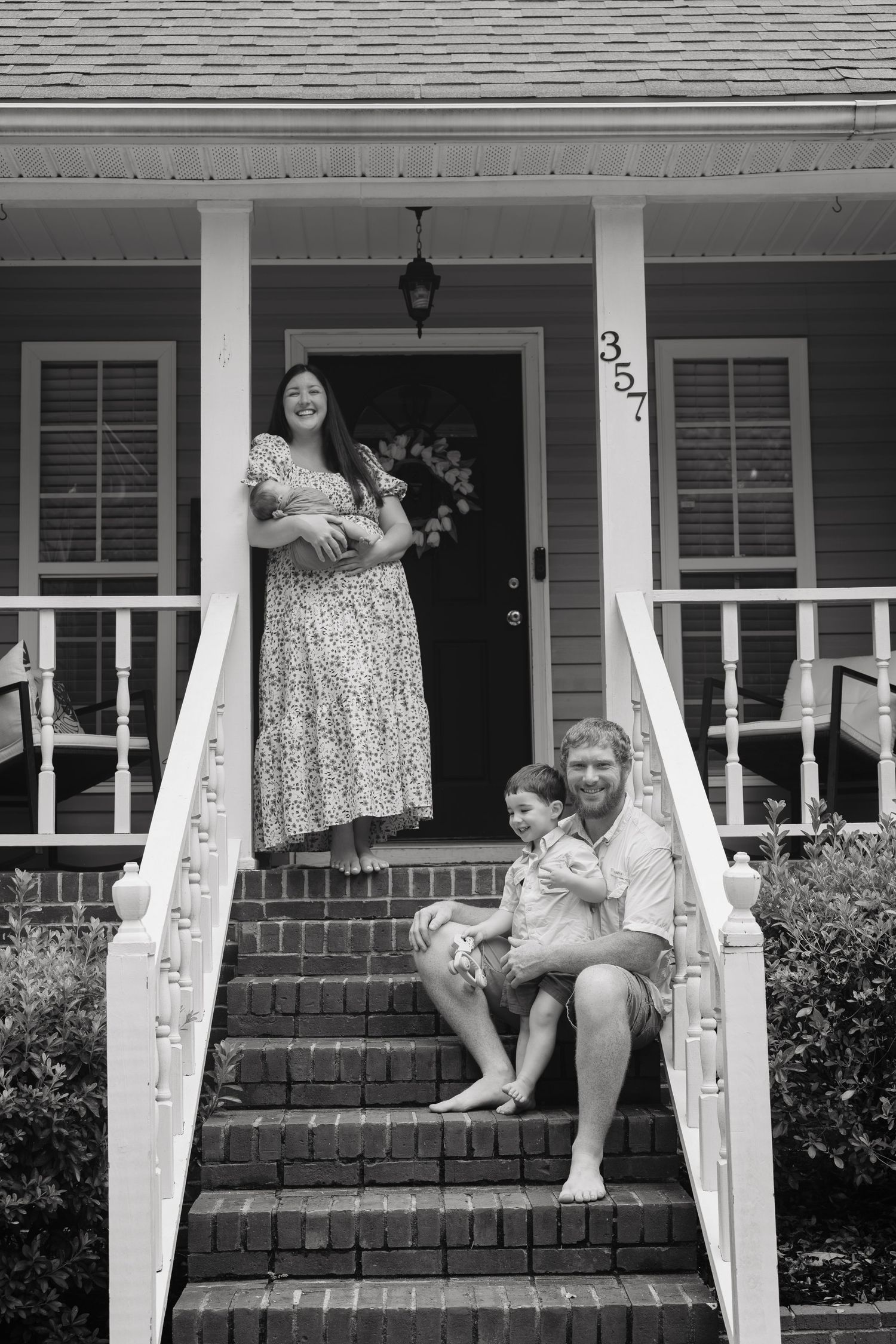 Black and white family portrait on brick porch steps, woman holding infant, man seated with toddler, house number 357.