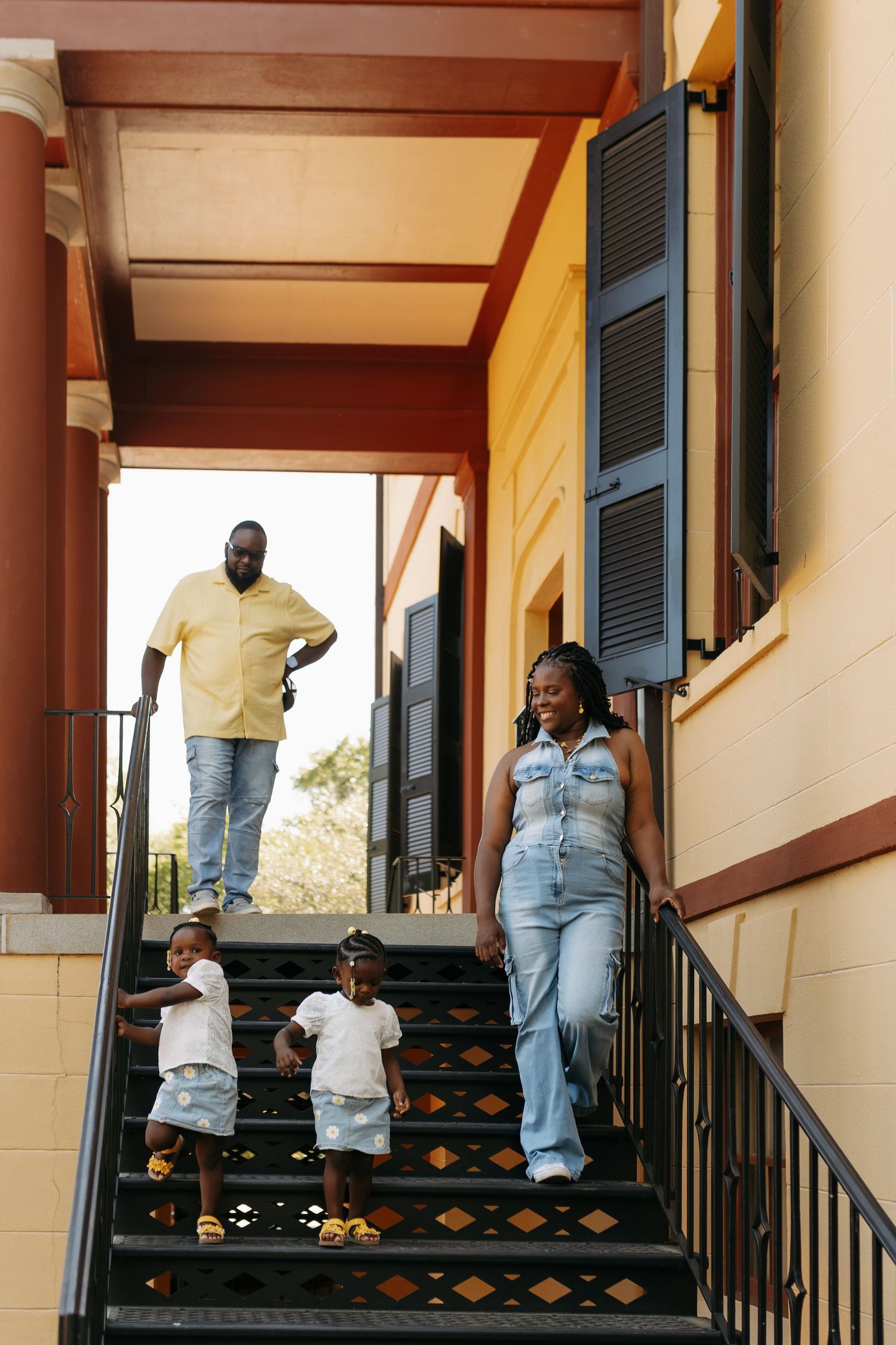 A joyful Black family with two toddlers walks down outdoor stairs of a colorful yellow and red building.