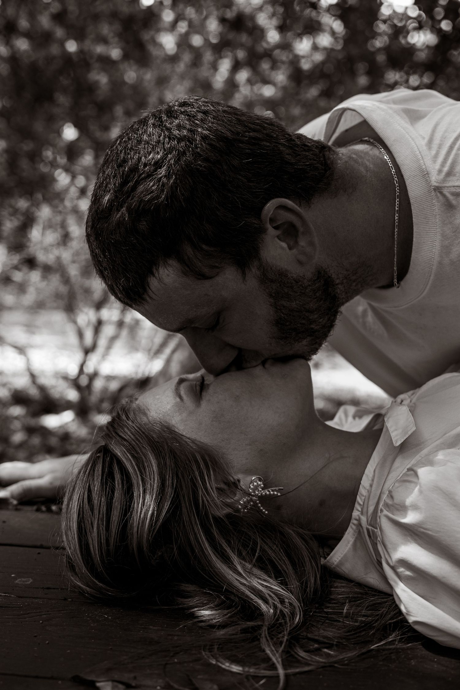 Black and white photo of a bearded man leaning down to kiss a woman lying on a wooden surface outdoors.