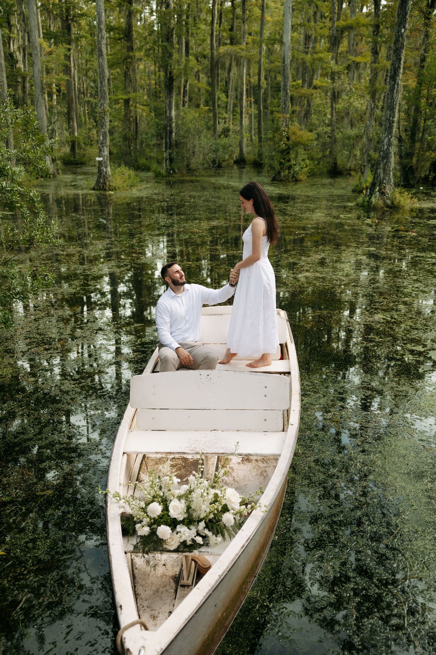 Couple in white attire sharing a romantic moment on a flower-adorned wooden boat in a lush green swamp.