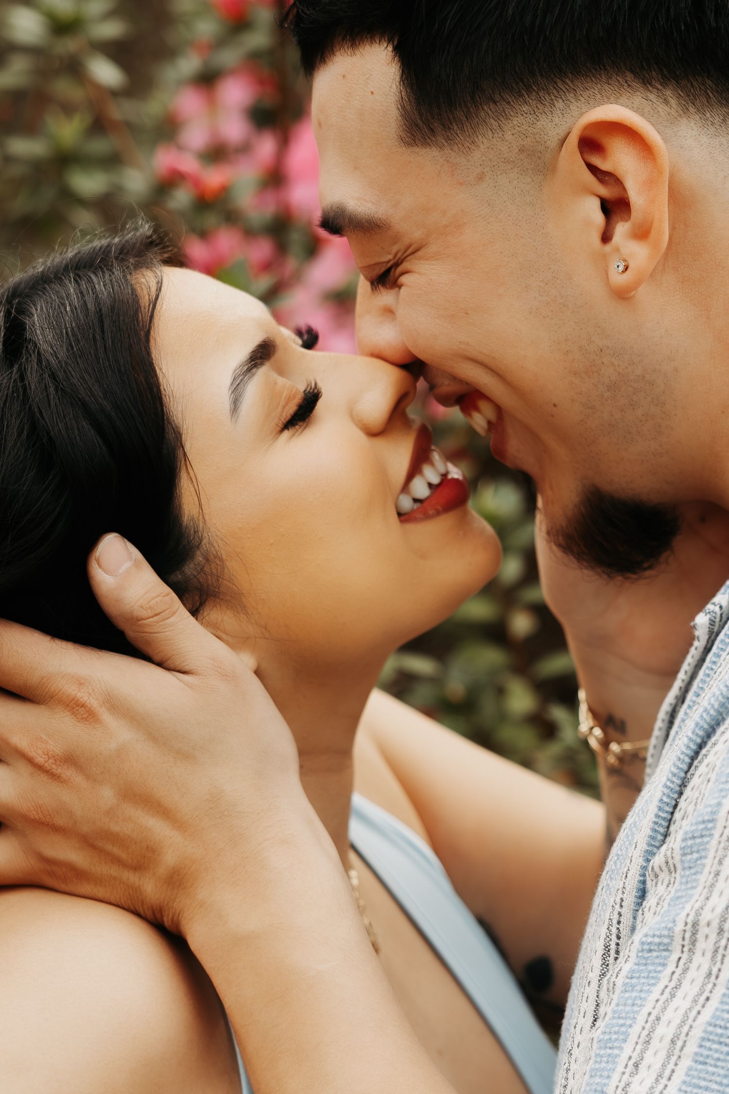 A smiling couple sharing a tender kiss outdoors with pink flowers in the background.