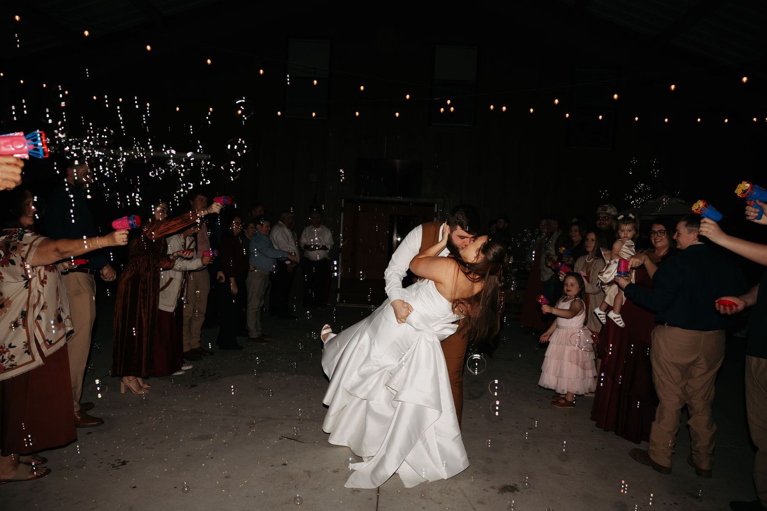 Bride and groom share romantic dip kiss on dance floor surrounded by wedding guests at outdoor evening reception.