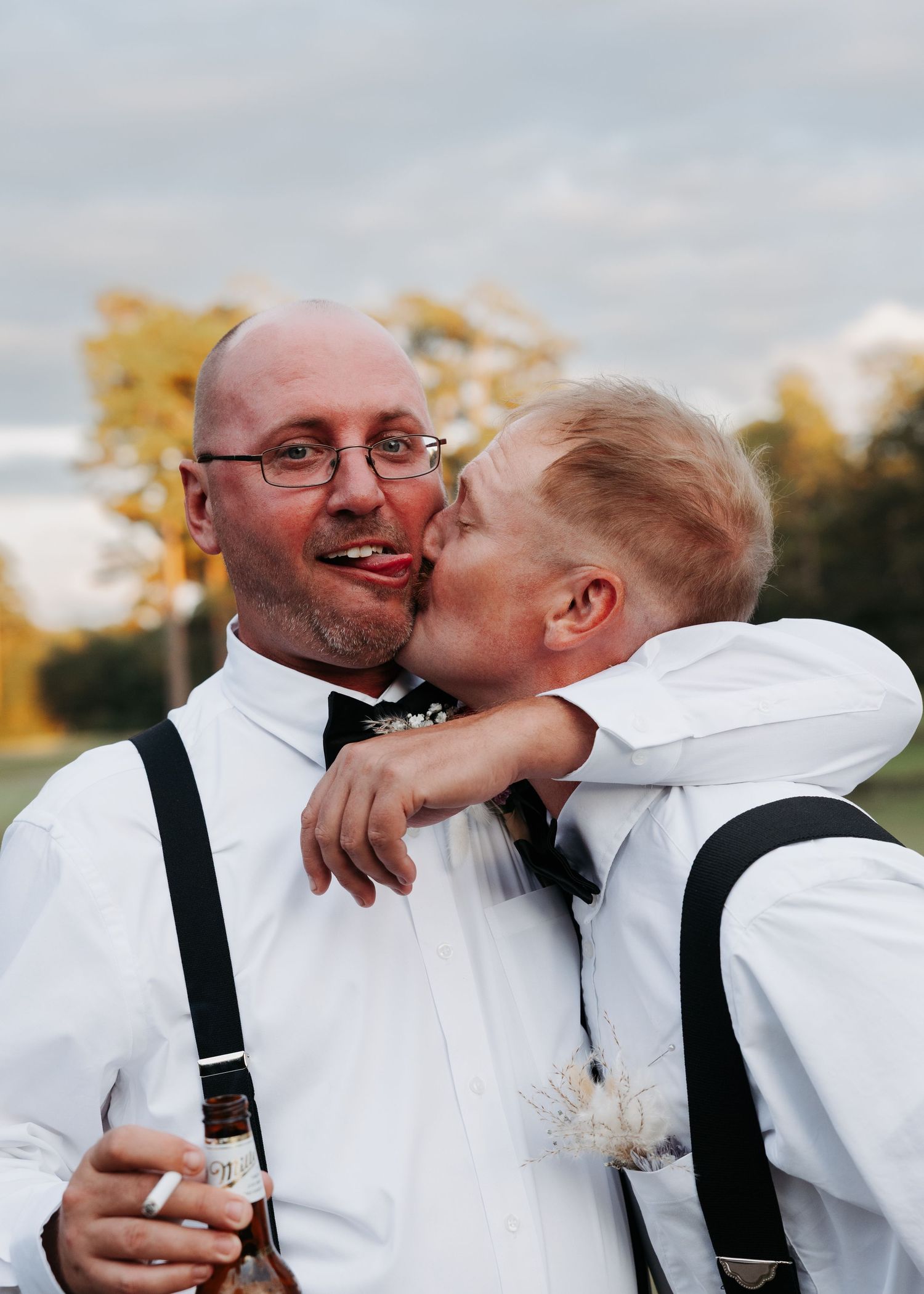 Two groomsmen in white shirts with suspenders sharing a joyful moment outdoors at a wedding reception.