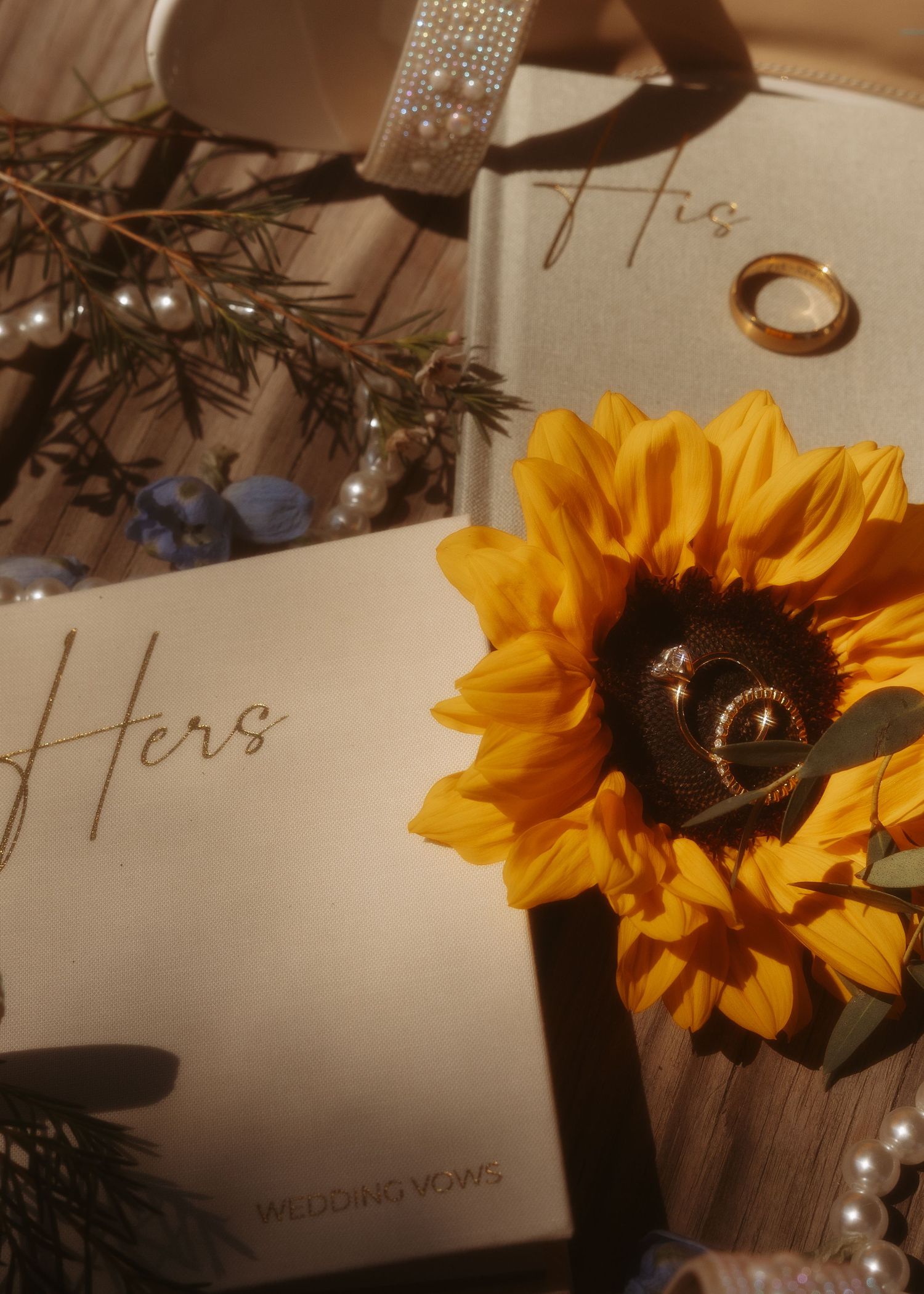 His and Hers wedding vow books with gold rings resting on a sunflower surrounded by pearls and greenery.