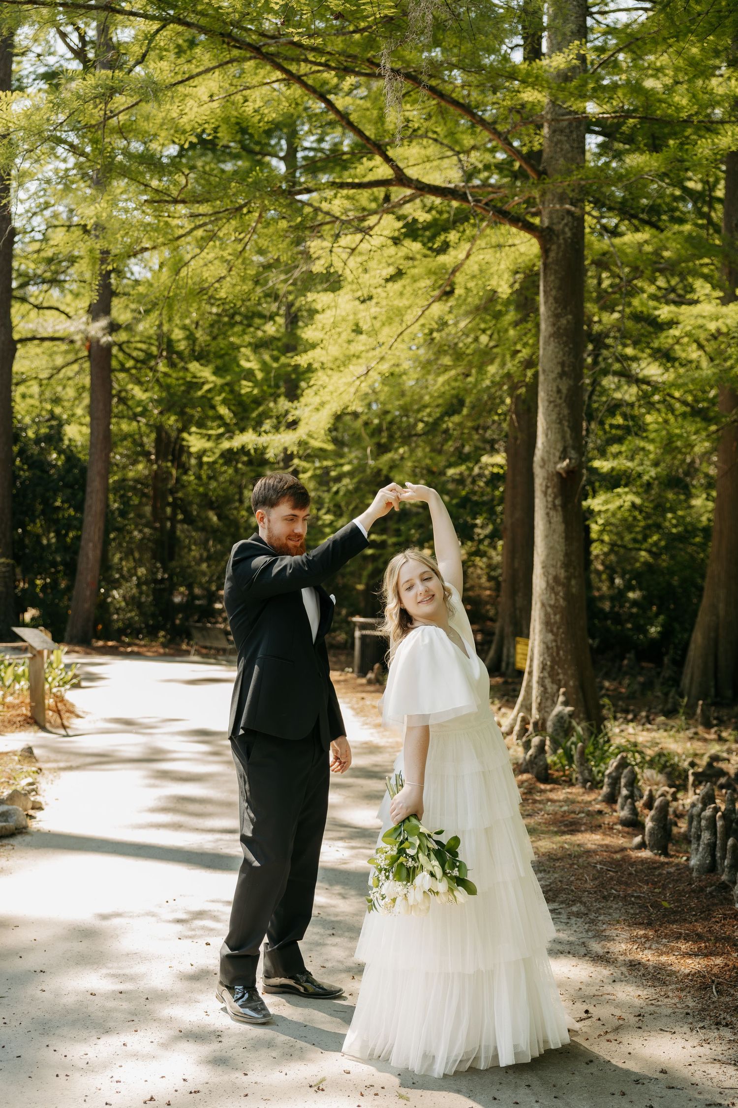 Groom twirling bride in white tiered dress on sunlit forest pathway.