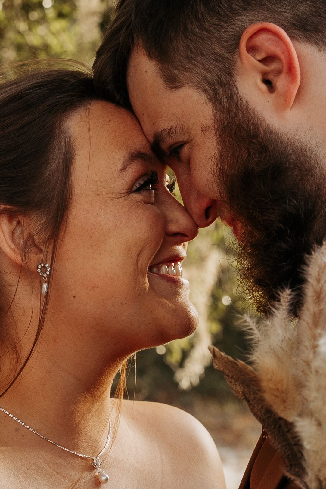 A smiling bride and bearded groom share an intimate forehead touch, nearly kissing outdoors in warm golden light.