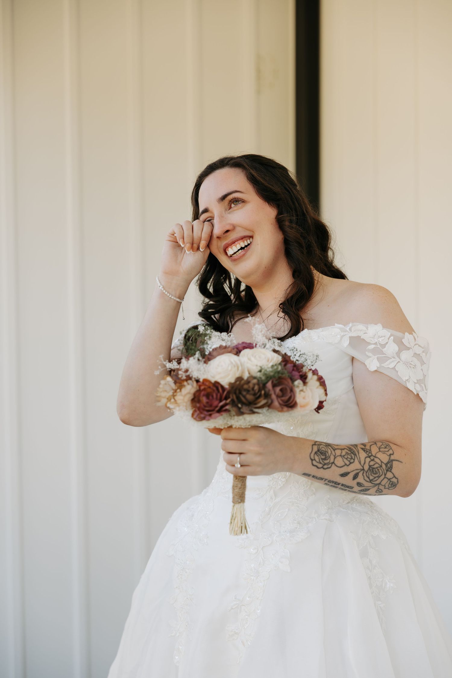Emotional bride in off-shoulder lace gown wiping tears, holding floral bouquet with tattoo visible, smiling joyfully.