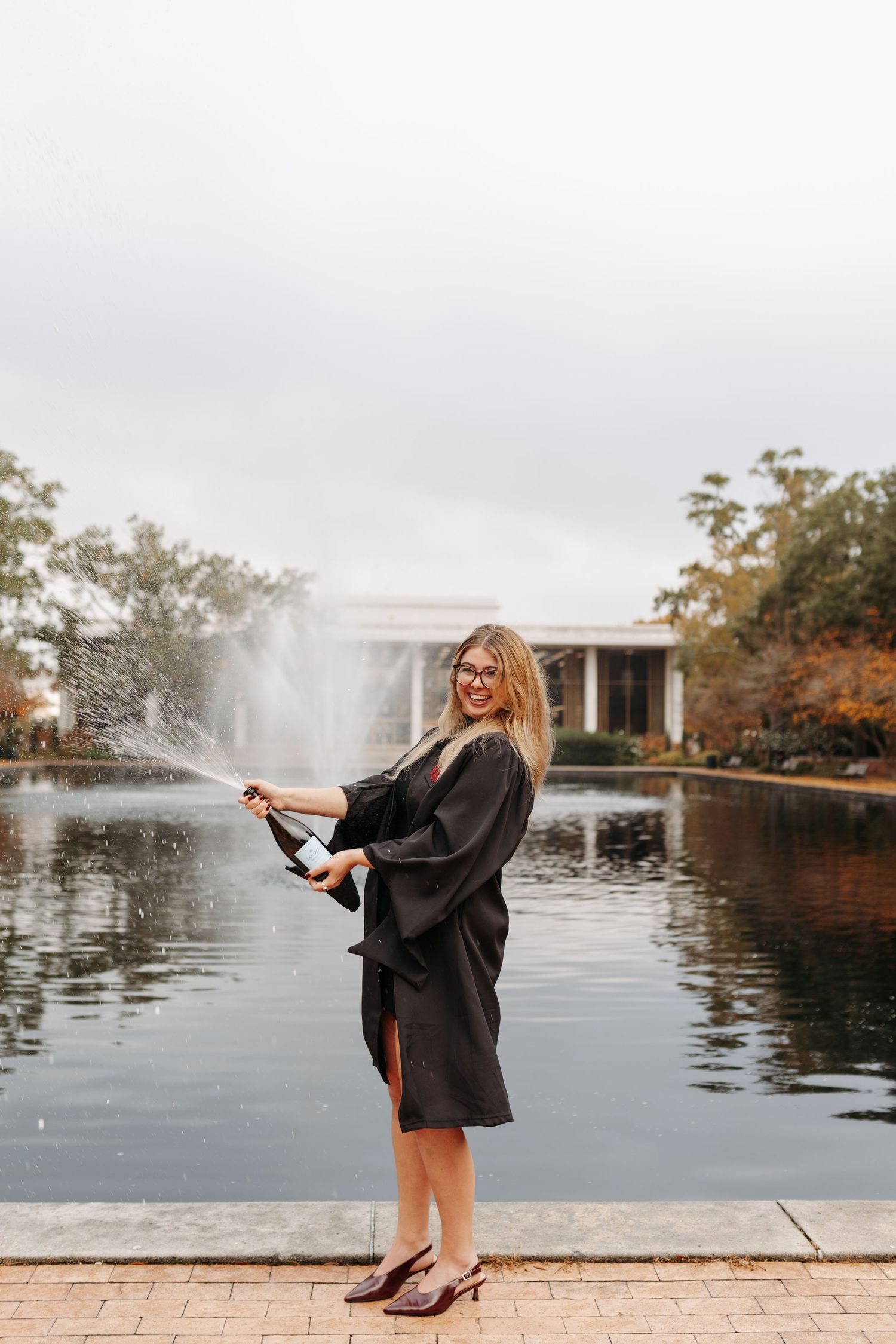 Graduate in black gown celebrates near fountain at sunset during graduation photoshoot.