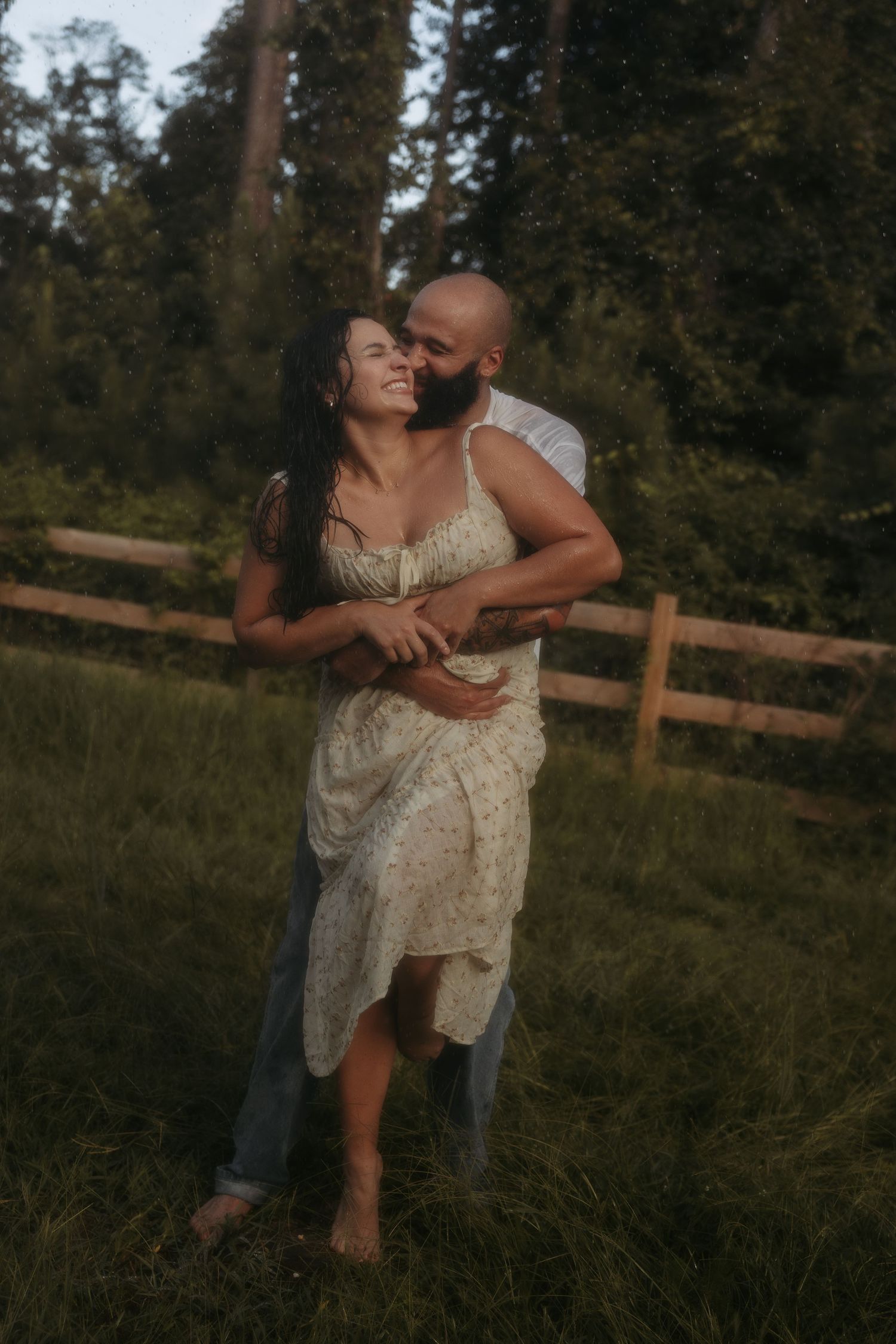 A couple embraces in a romantic moment on grassy field with rustic fence and trees in background at sunset.