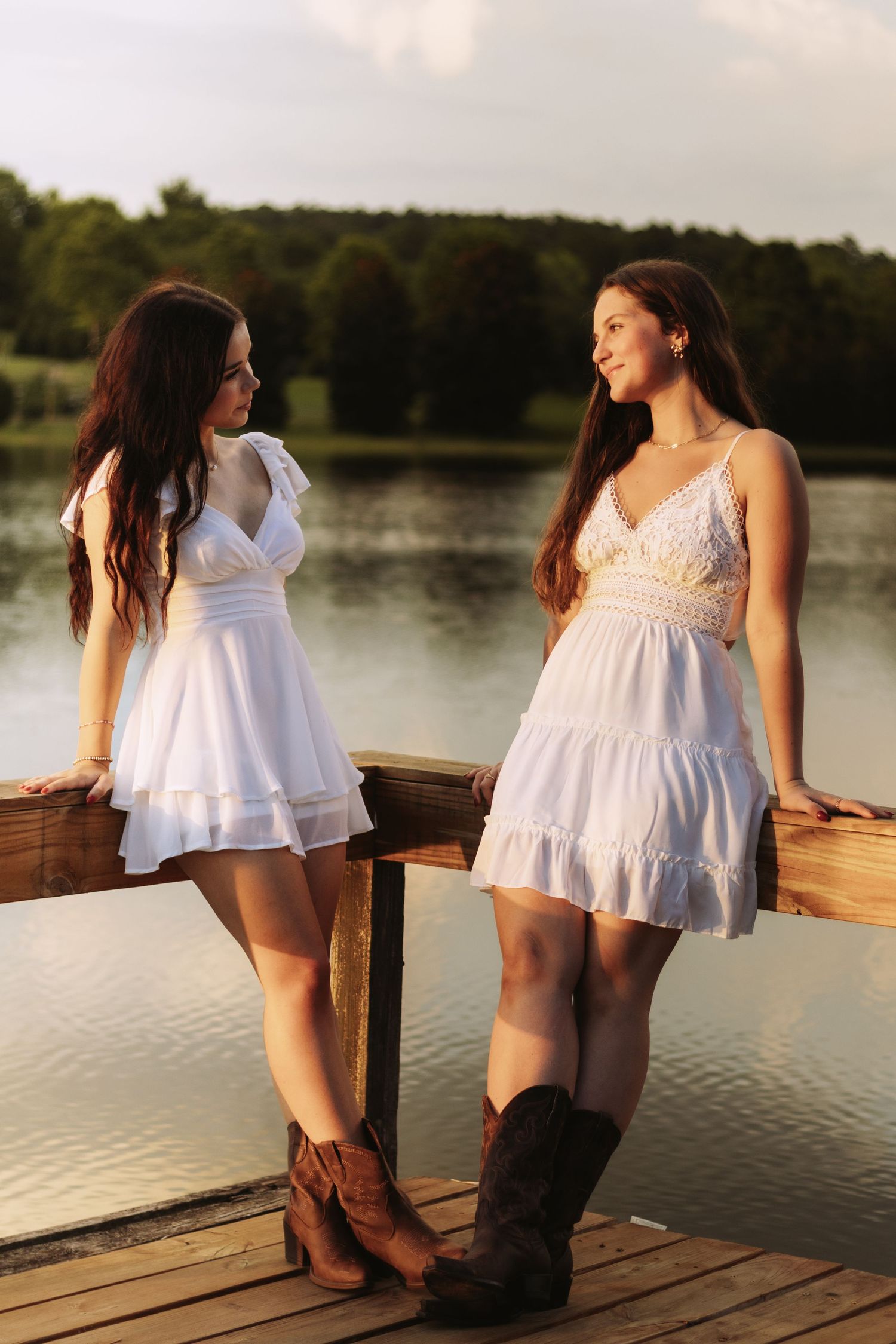 Two women in white summer dresses and cowboy boots stand together on a wooden dock by a lake at sunset.
