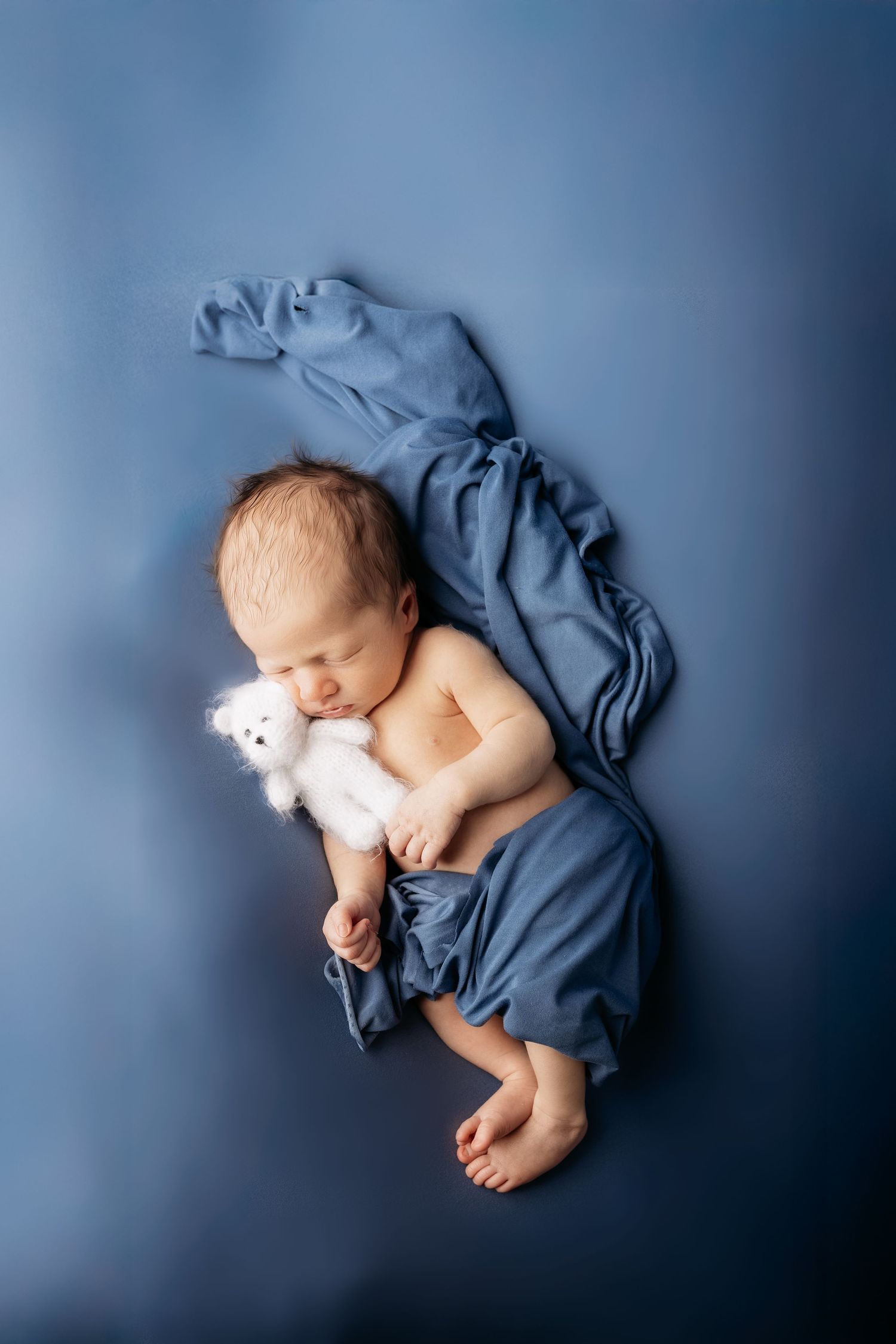 Newborn baby sleeping peacefully on blue fabric, cuddling a small white teddy bear.