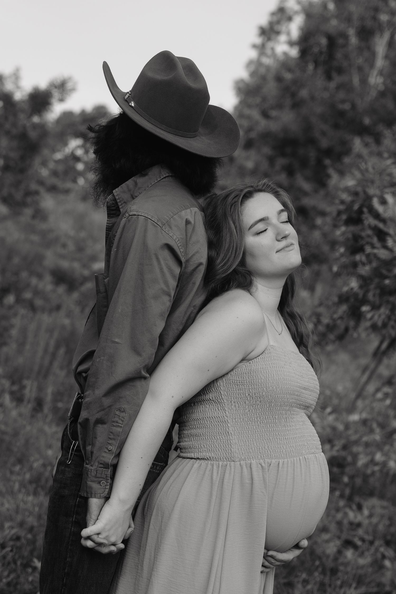Black and white maternity photo of pregnant woman in dress holding hands with partner in cowboy hat outdoors.