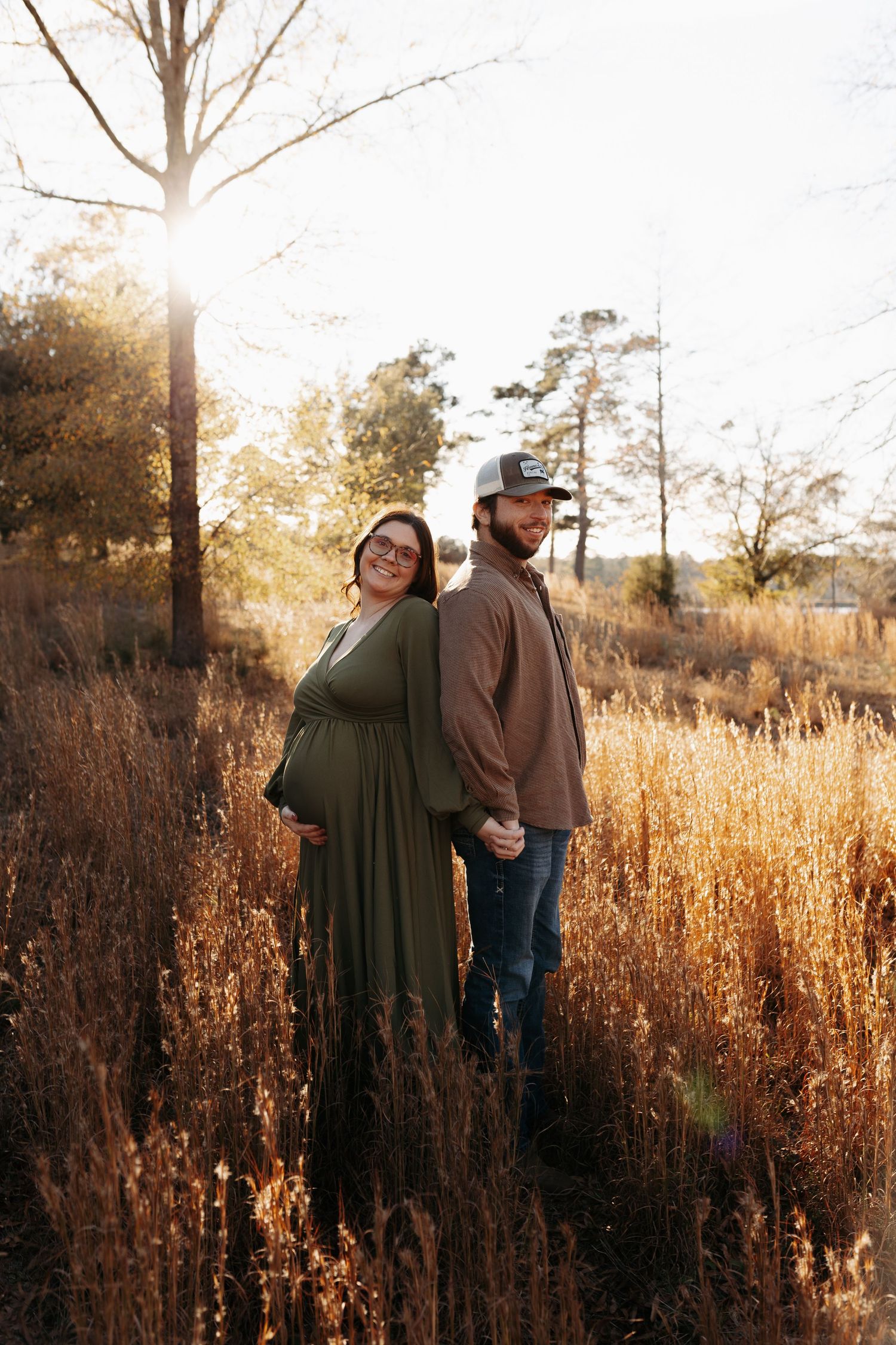 A couple stands together in a golden field of tall grass at sunset with sunlight streaming through bare trees.