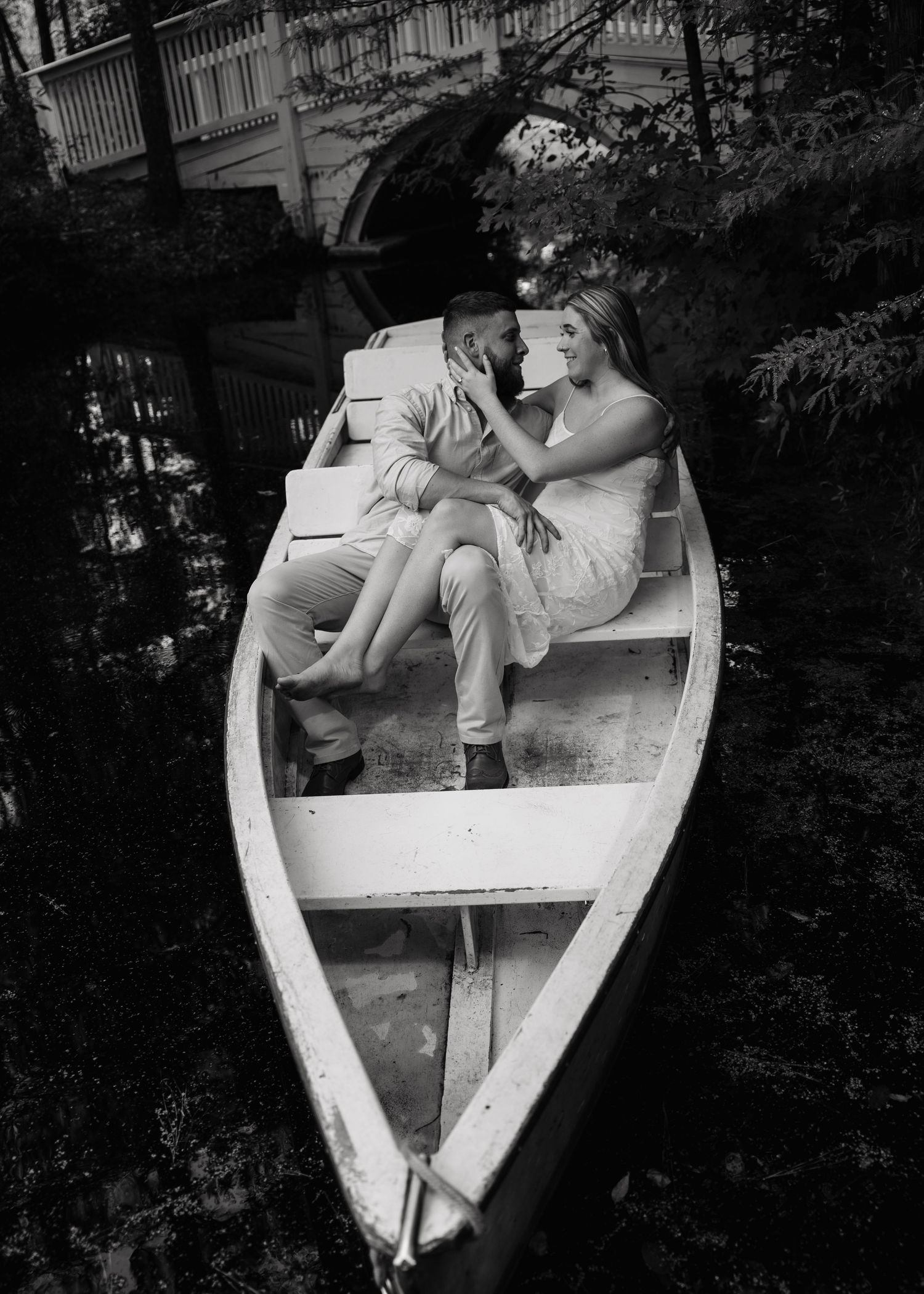Black and white photo of a romantic couple sitting close together in a white rowboat surrounded by lush trees.