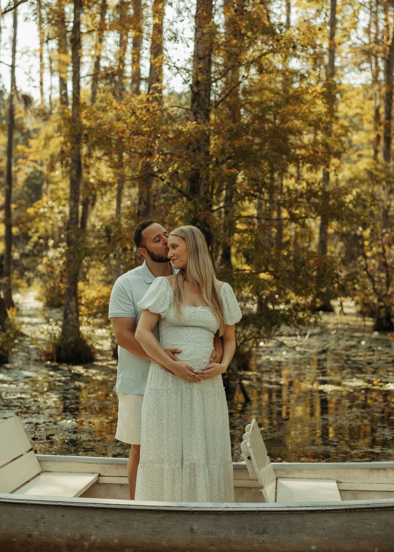 Couple standing in a boat surrounded by autumn trees, man kissing pregnant woman's head while she smiles.