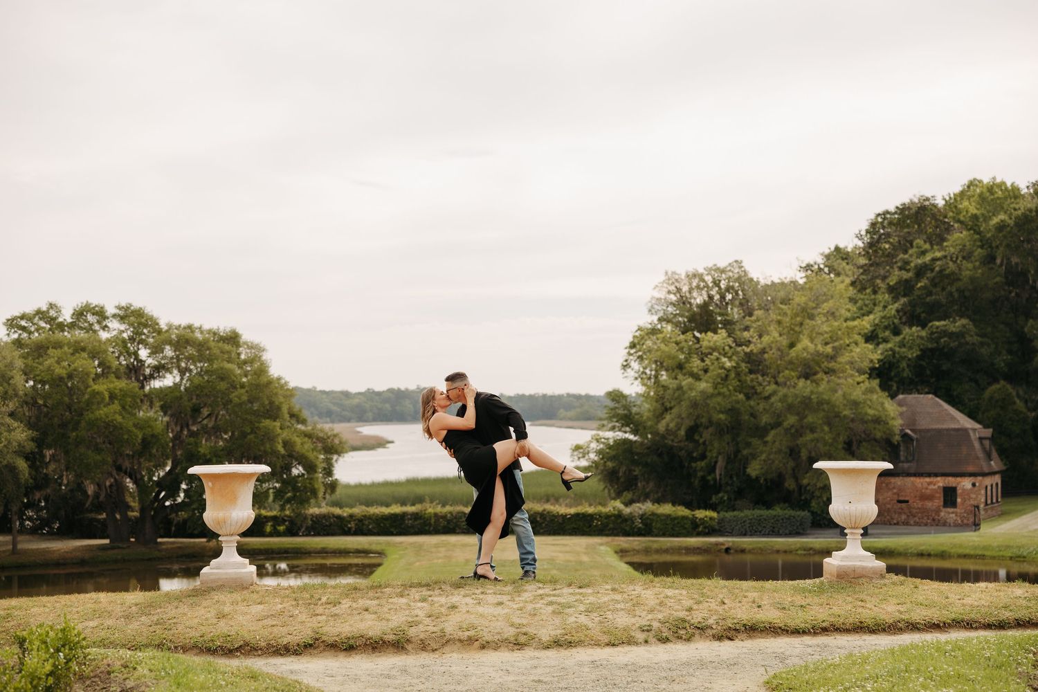 Couple shares romantic lift pose in formal garden with water view, white urns, and lush trees in background.