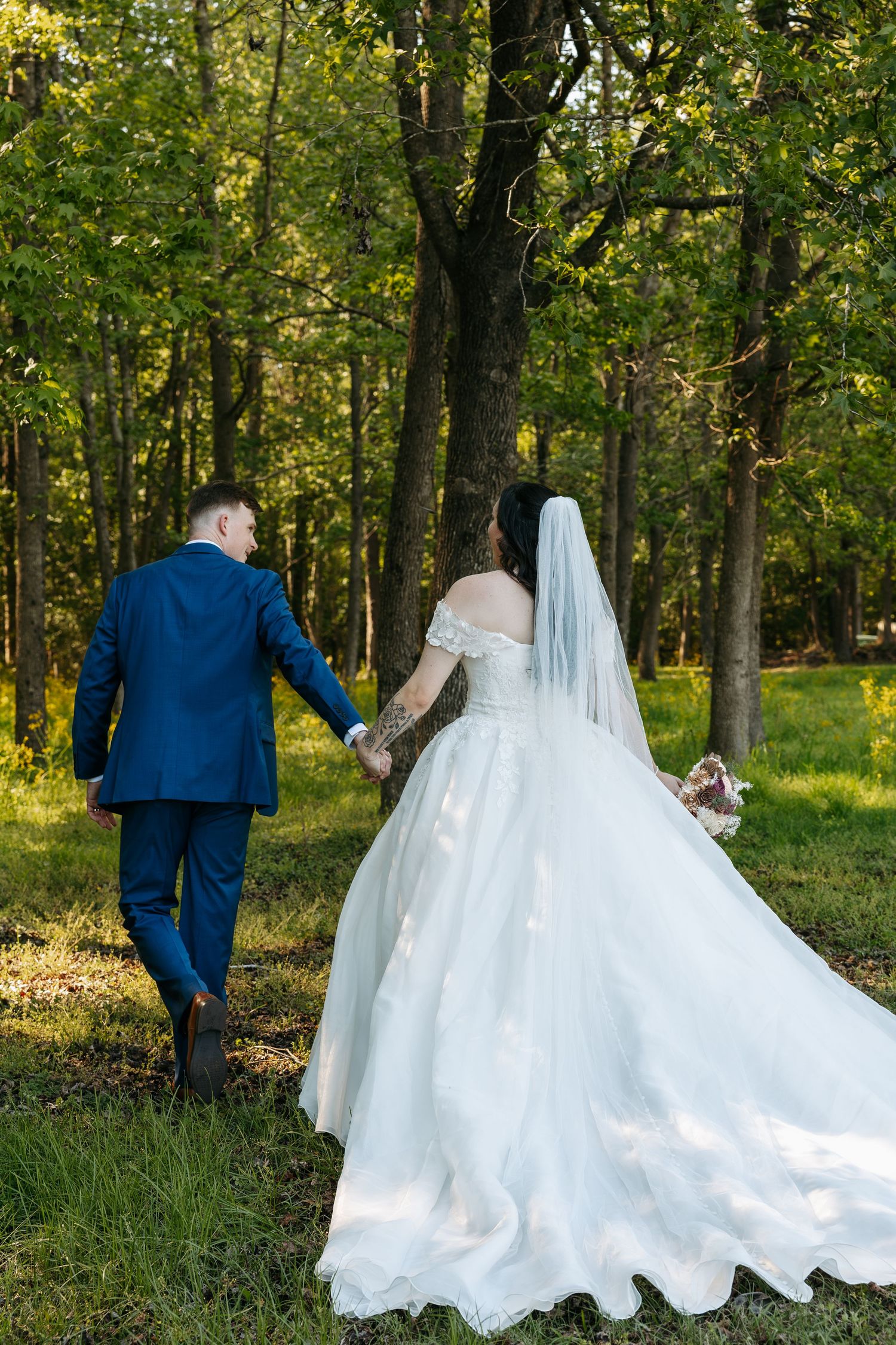 Bride and groom walk hand in hand through lush green forest, bride's gown trailing behind them.
