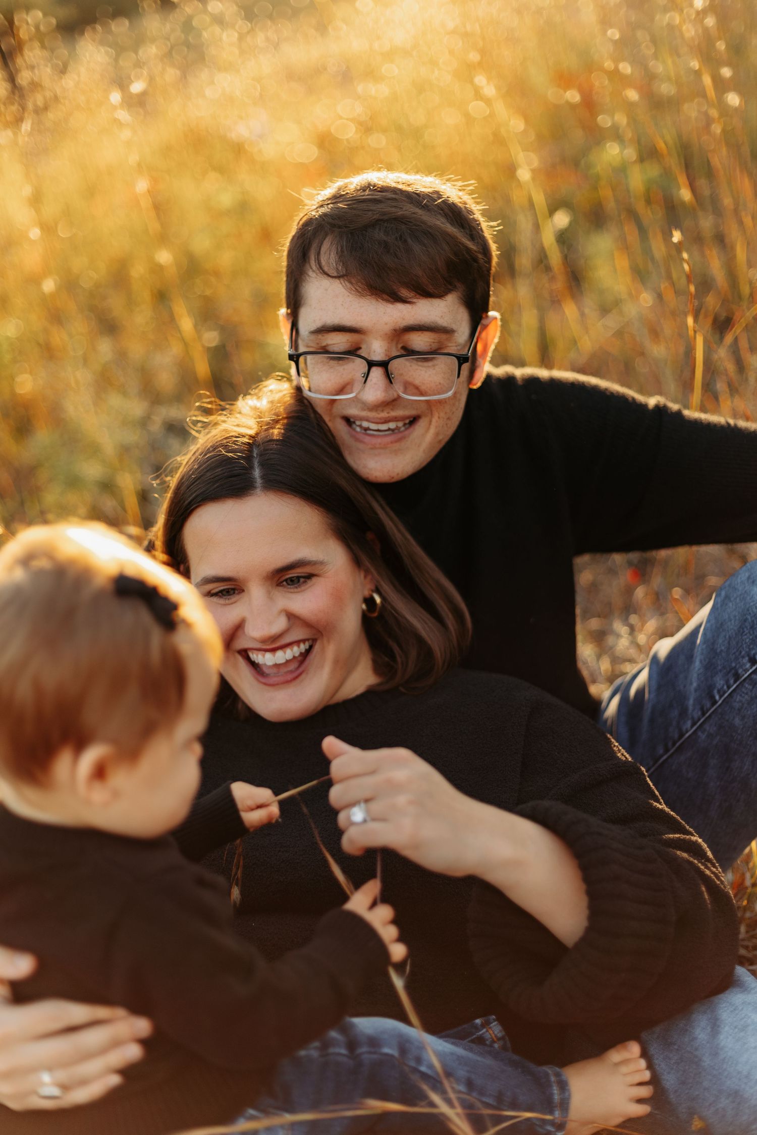 Family laughing together outdoors in golden autumn field, two adults embracing child in warm fall sunlight.