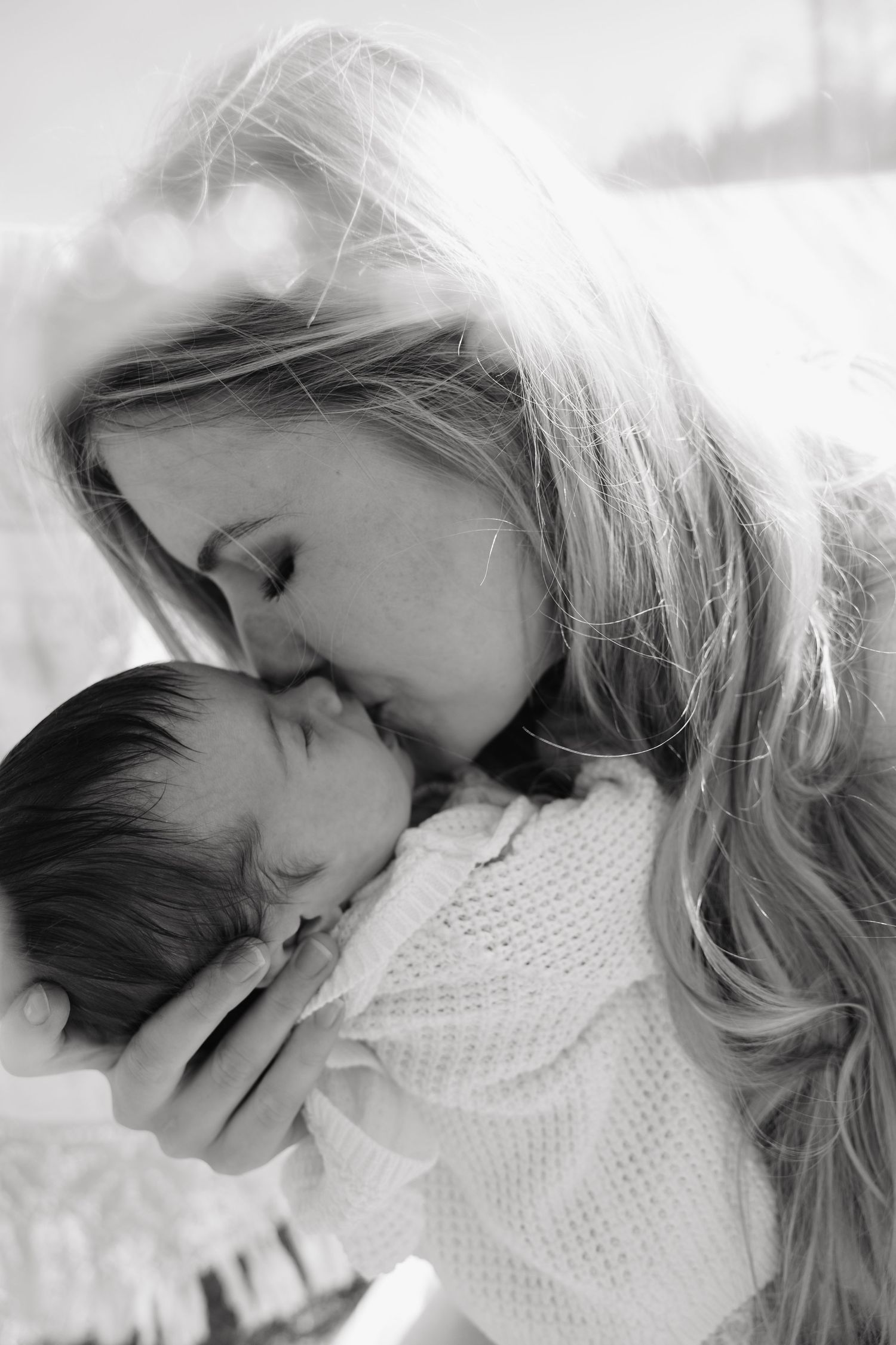 Black and white photo of a loving mother gently kissing her sleeping newborn baby.