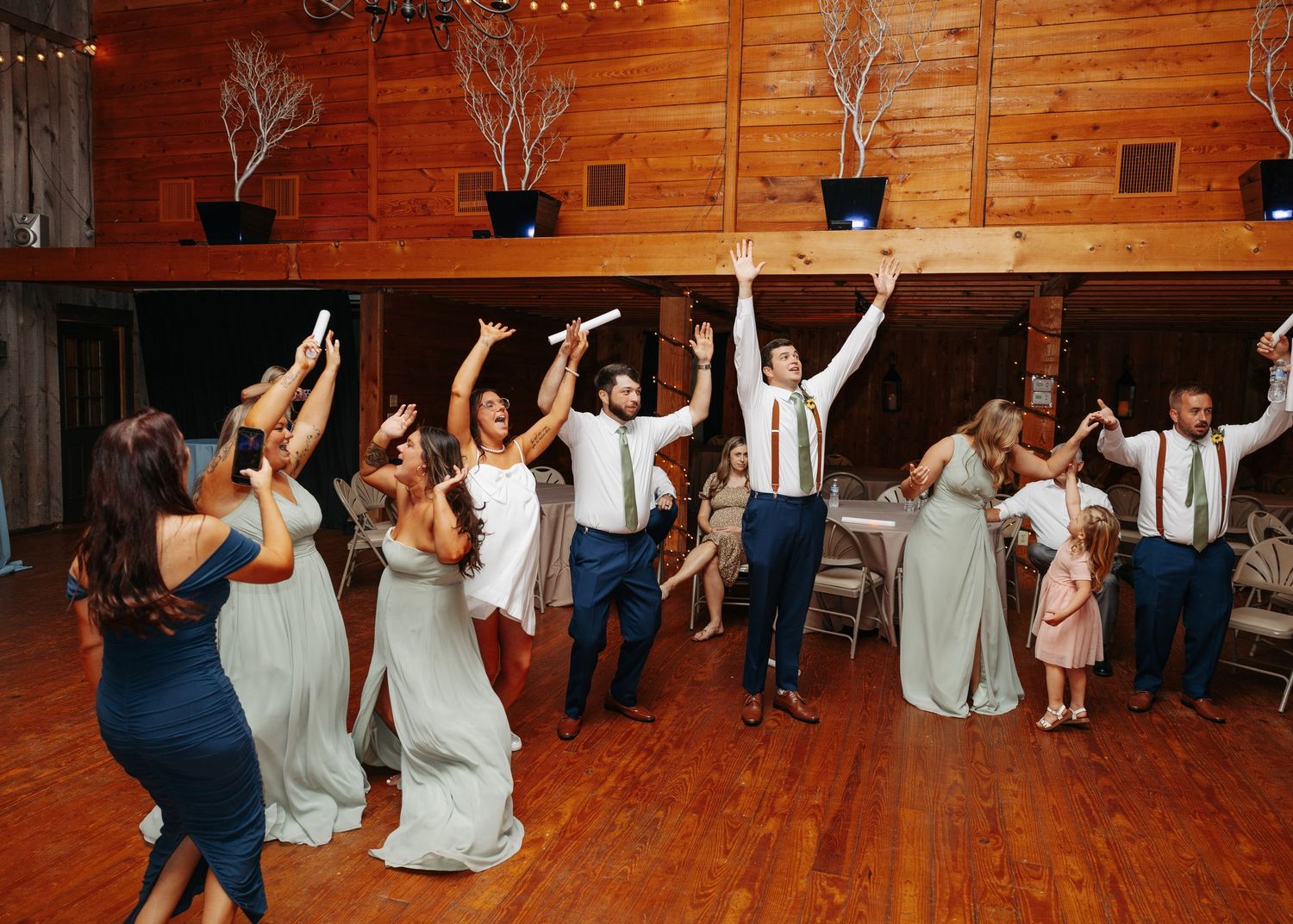 Wedding party dancing joyfully on wooden dance floor in rustic venue with raised hands during reception celebration.