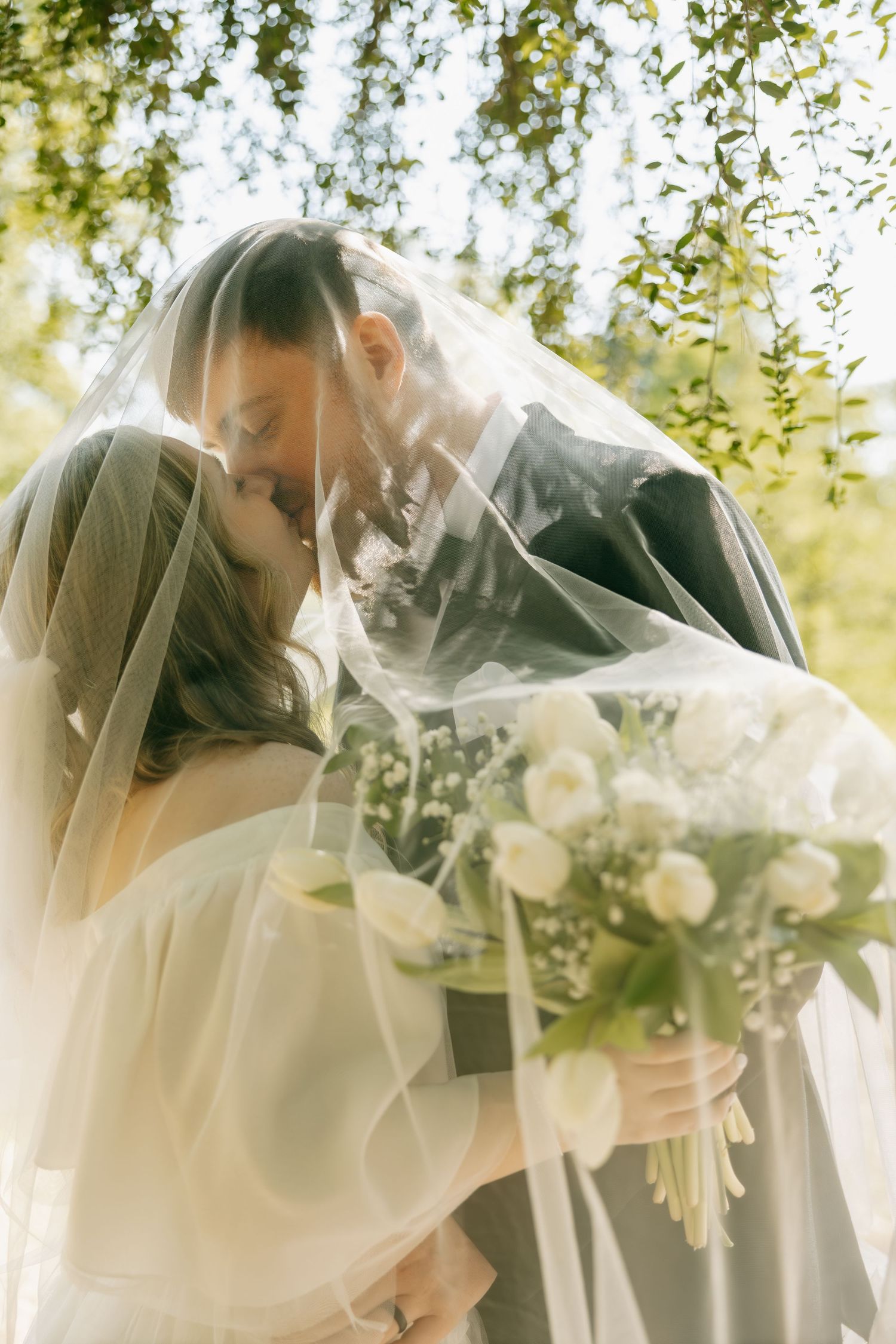 Bride and groom sharing a romantic kiss under a flowing veil, with a white rose bouquet and lush green trees.